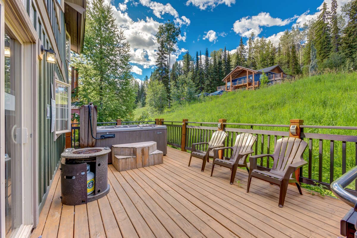 Picture of an outdoor area in the rocky mountains in Canada, containing a hot tub, deck chairs and fire pit. 