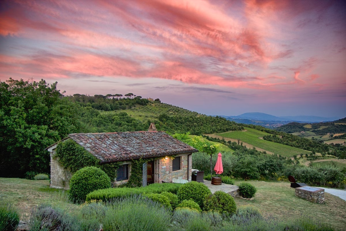 A small stone cottage sits in the foreground with a countryside vista and pink and purple sunset in the background