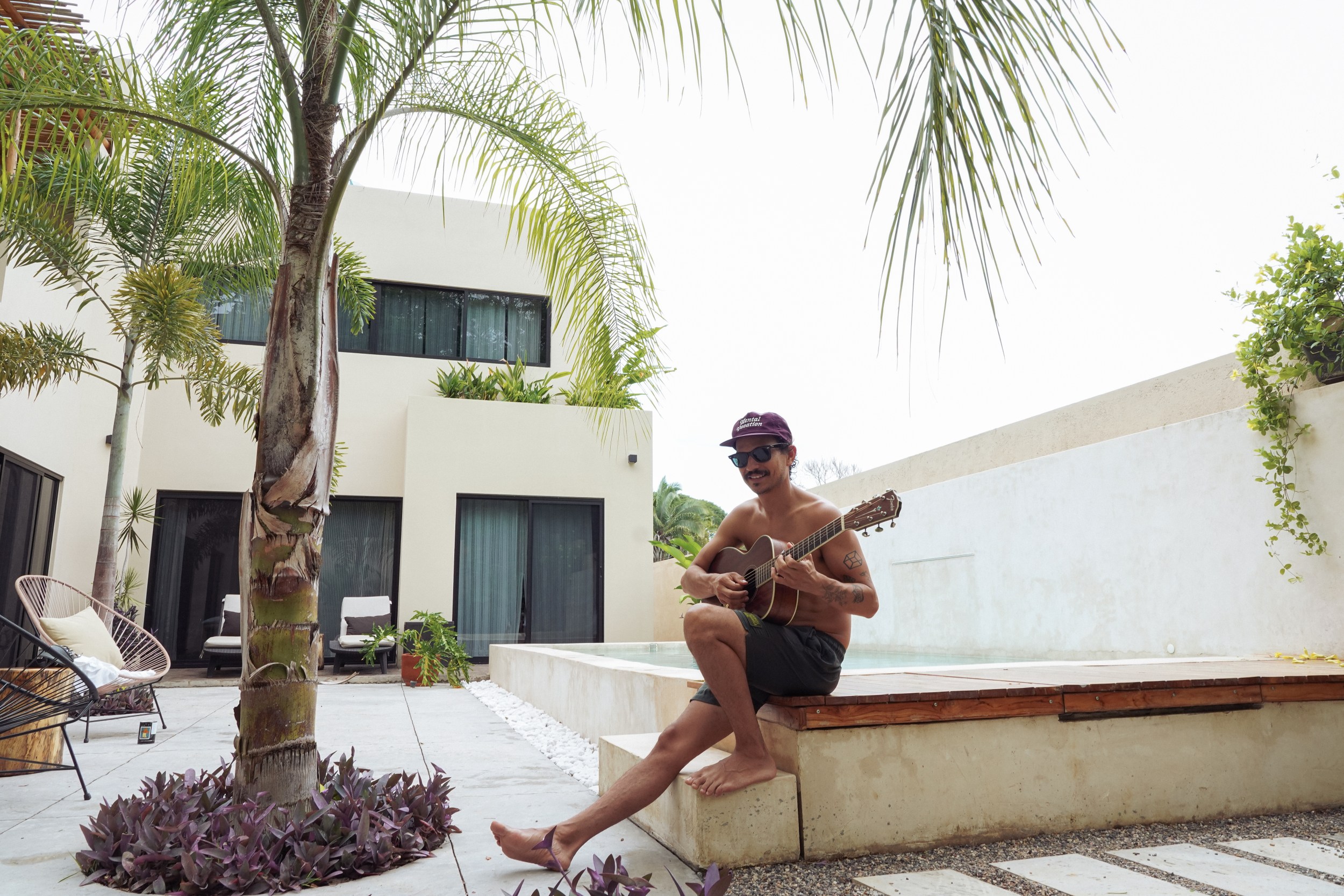Caloncho sentado en la orilla de la alberca bajo una palmera sosteniendo una guitarra