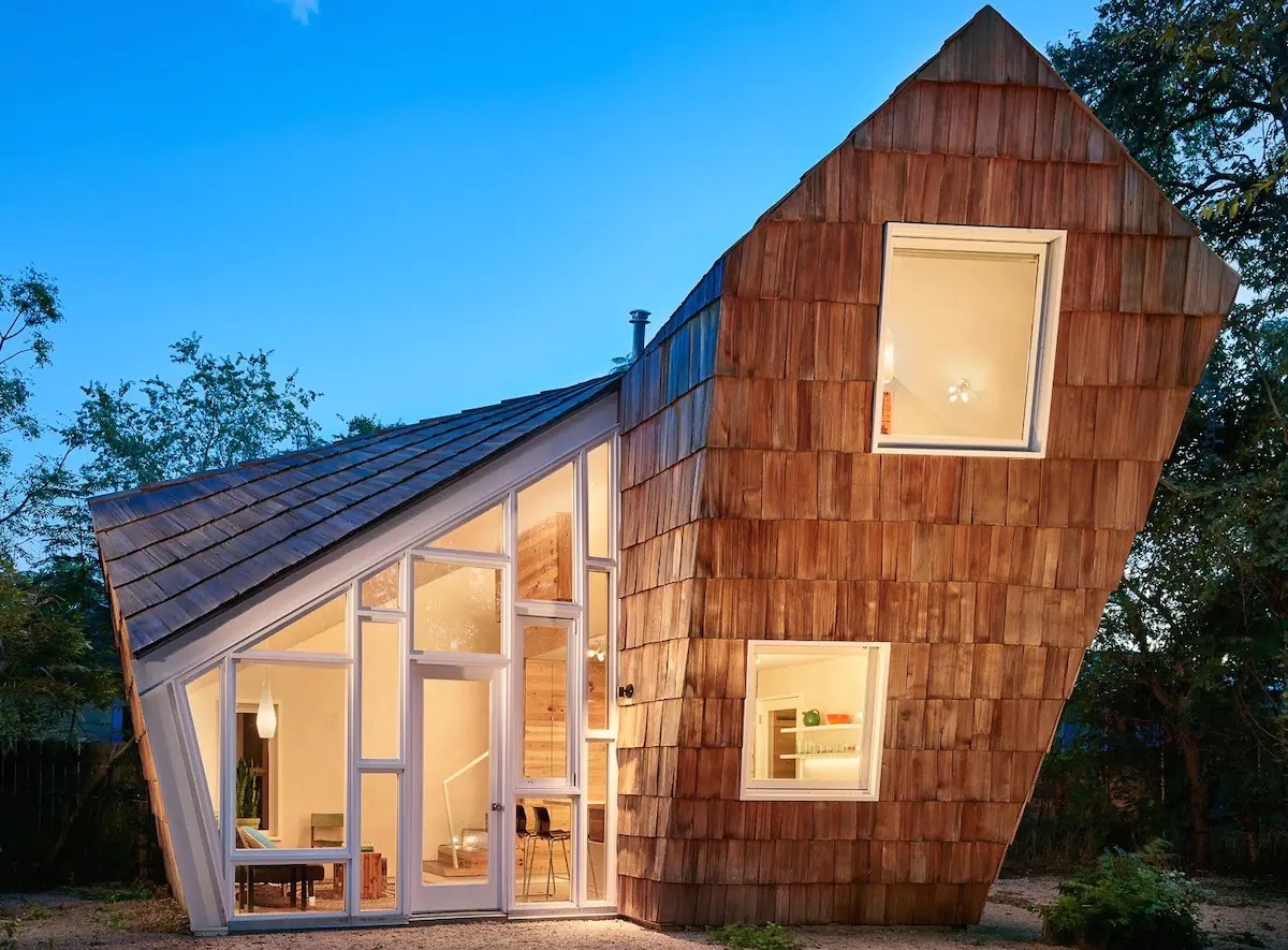 A wood paneled angular house with lights illuminating through large windows at dusk. 