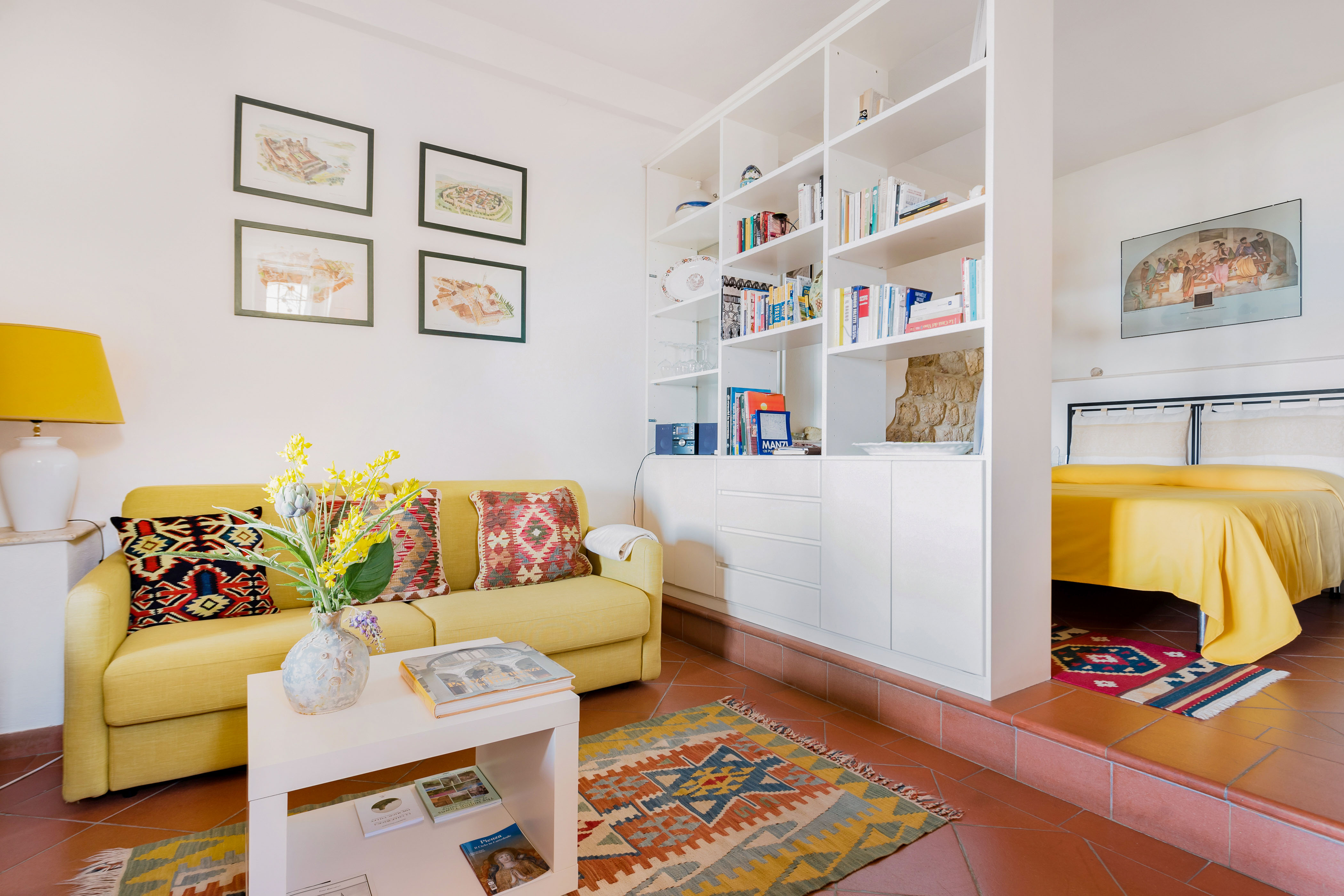 Sunken living room with a yellow couch and large white bookcase that steps up to a bedroom.