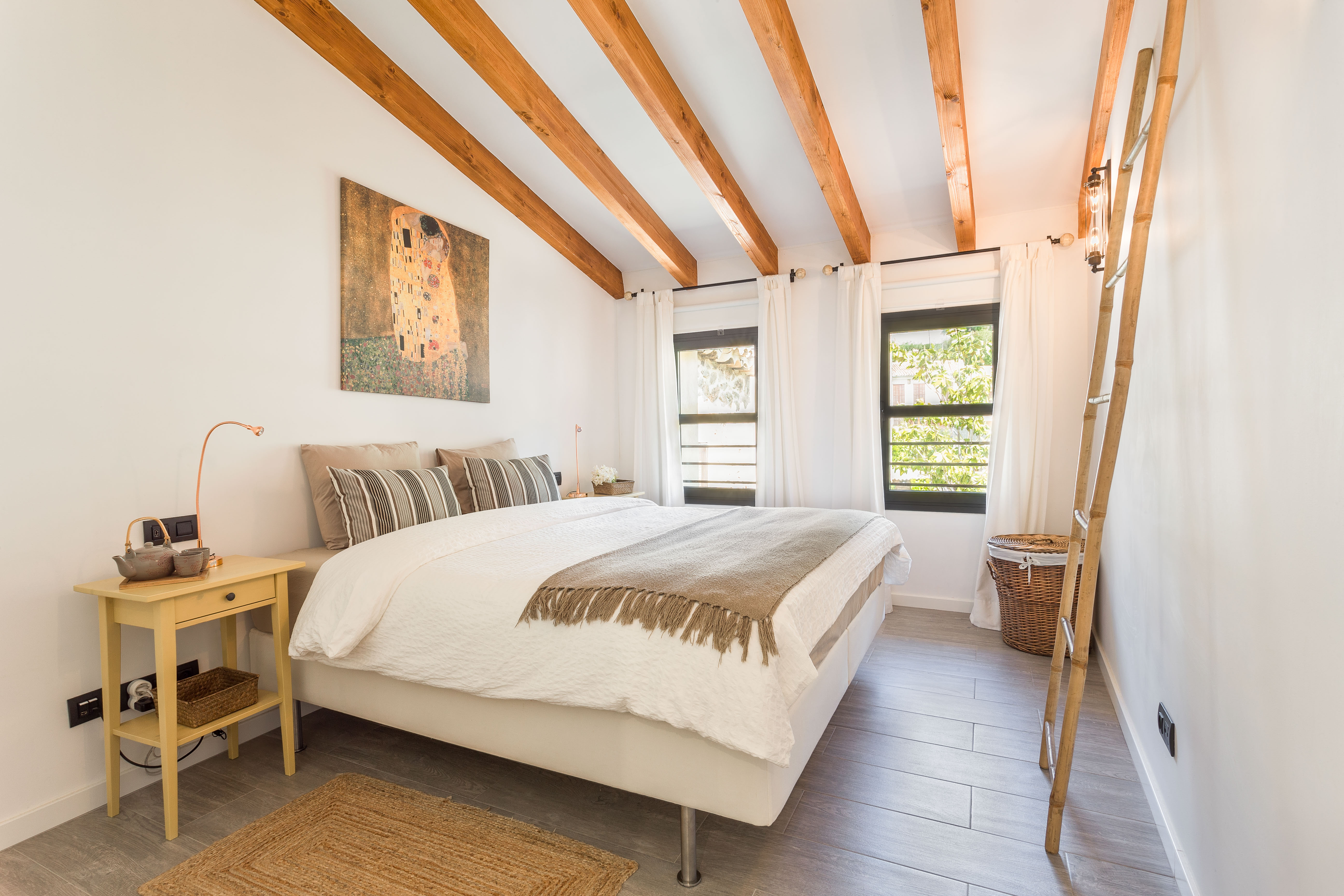 White bedroom with wood beamed ceiling and a decorative wooden ladder lit by two large windows.