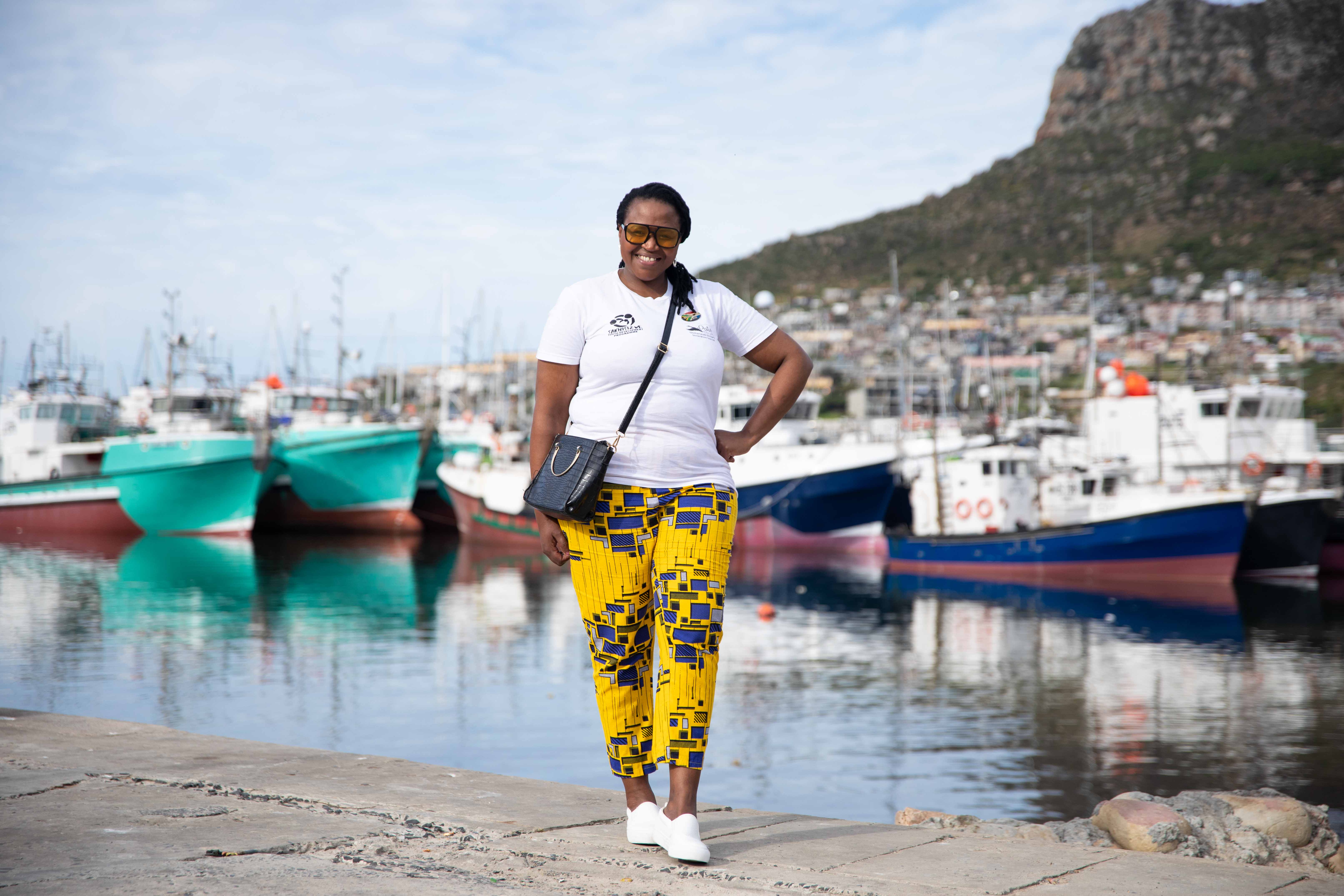 A woman in a white tshirt, yellow patterned pants and a black purse stands smiling in front of a boat dock. 