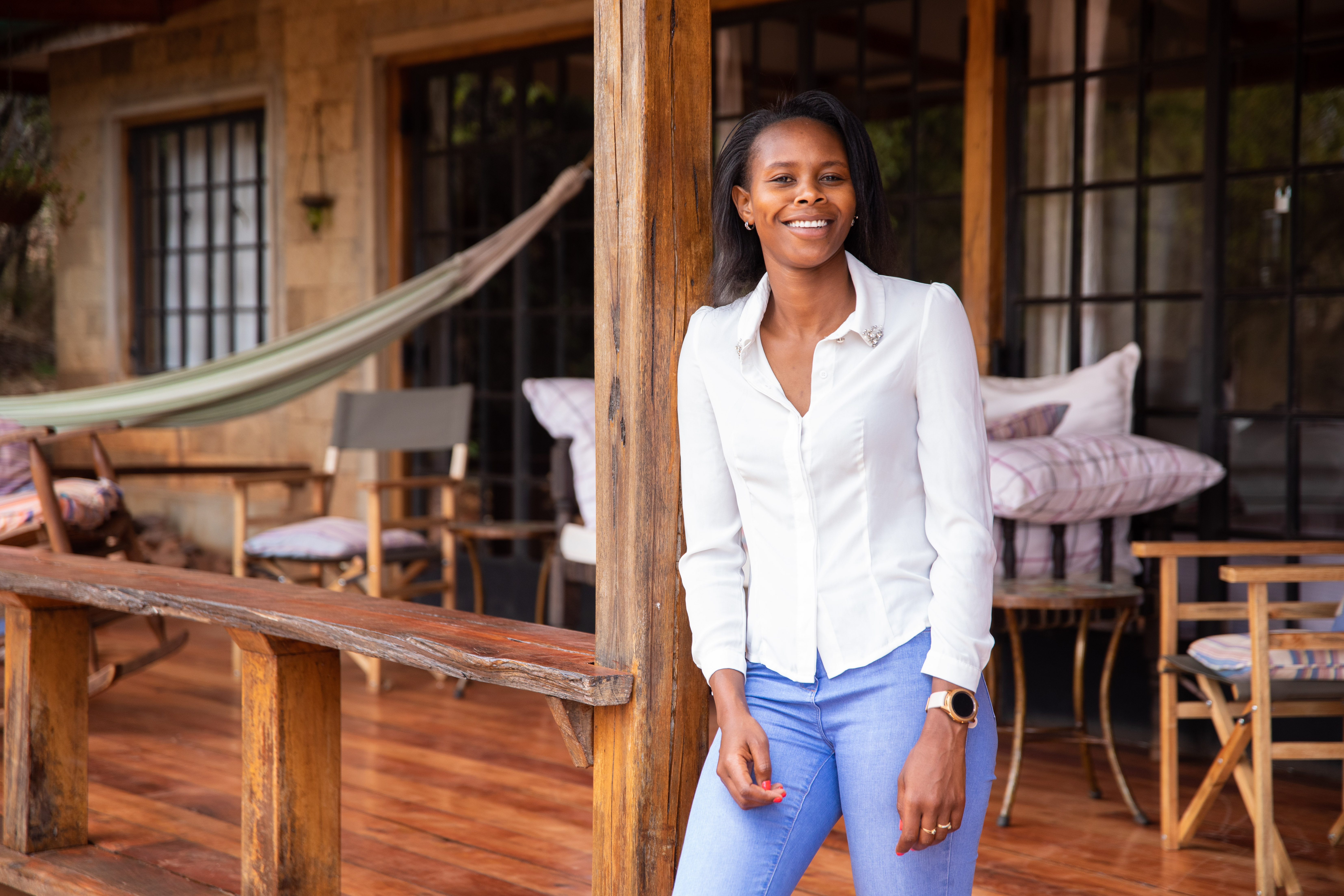 A woman in a white shirt and blue jeans leans against a wooden porch pillar, smiling with outdoor furniture behind her.