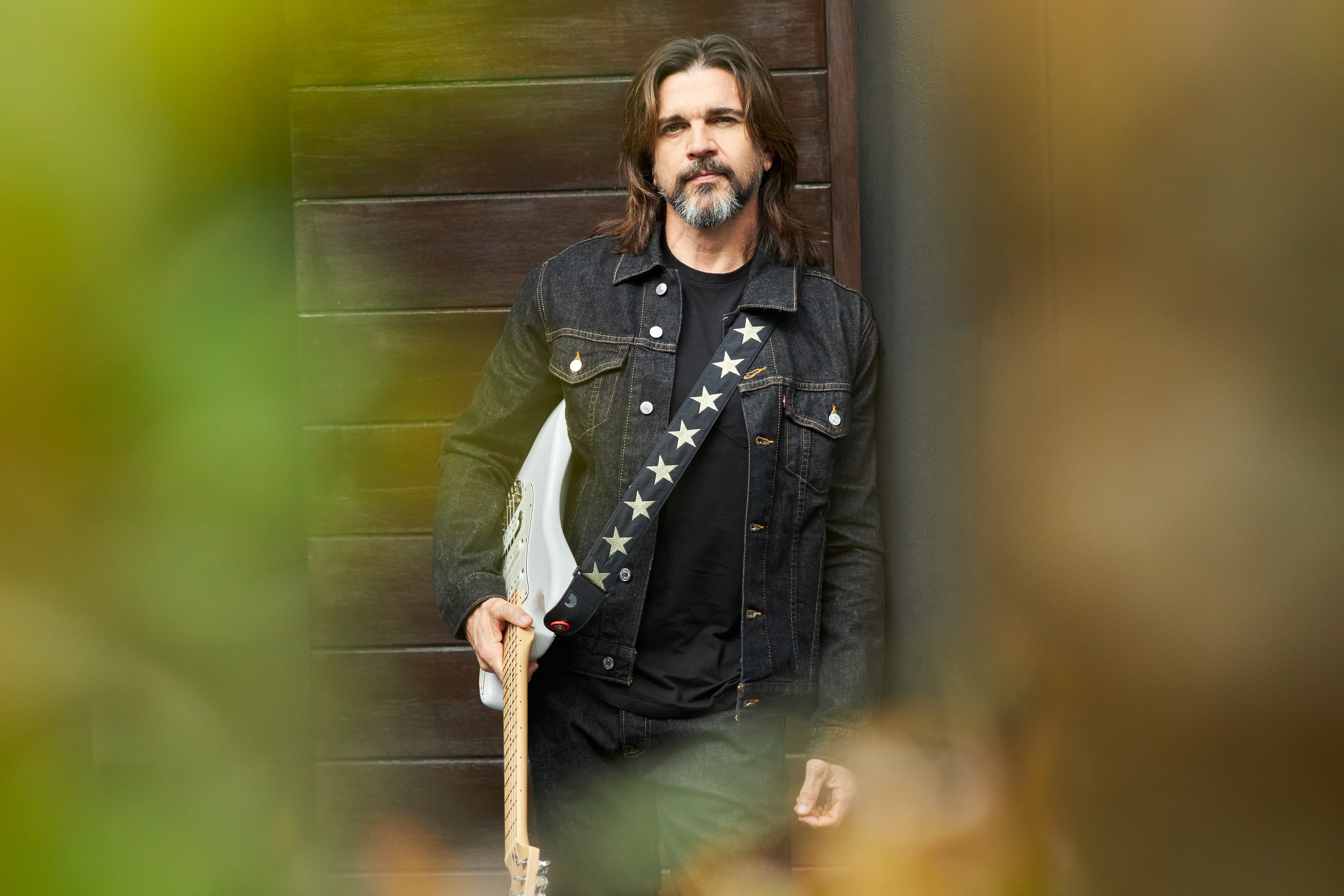Guitar player with long hair, a mustache and black leather jacket leans against a wall with his white guitar slung around him.