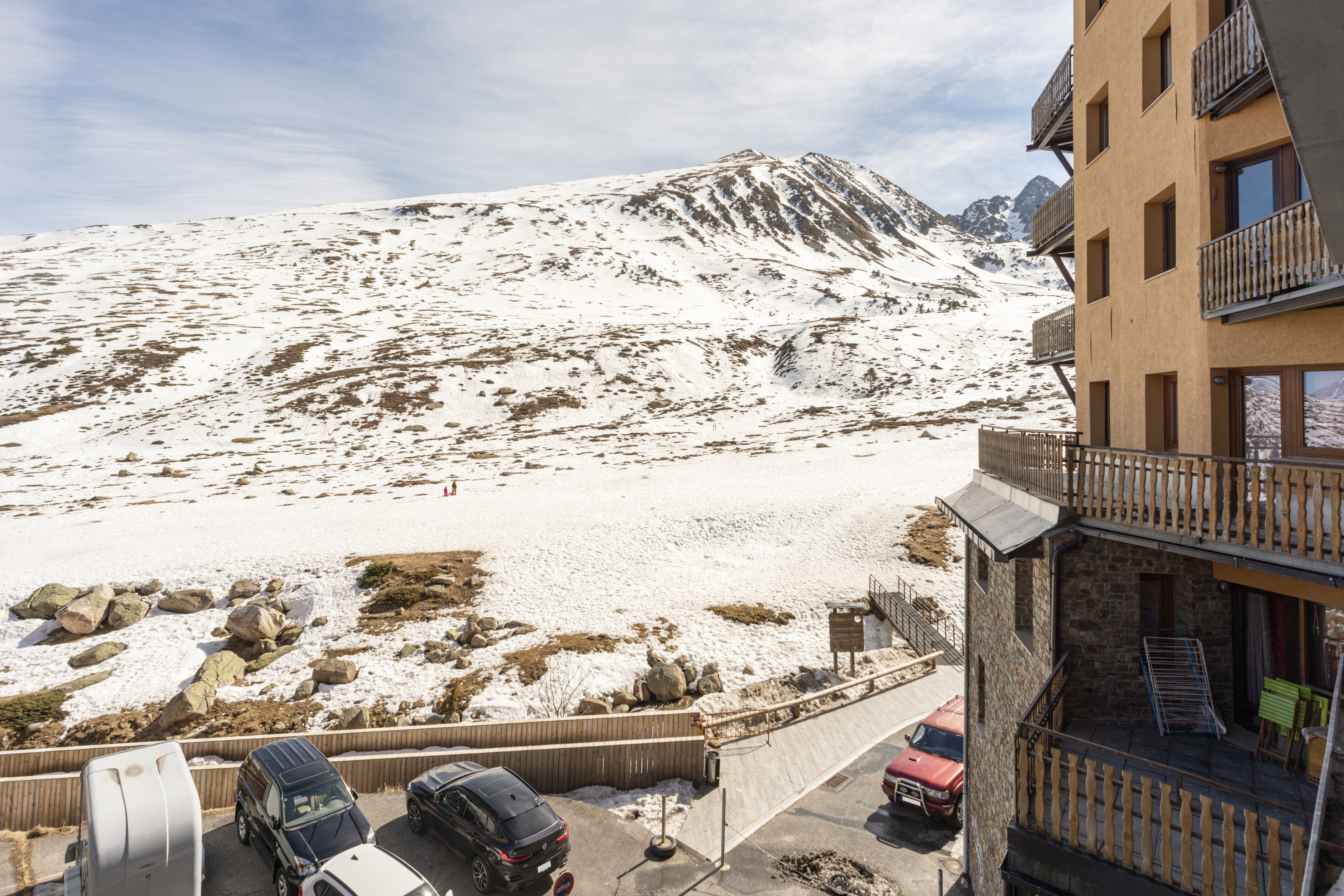 View to a mountain with snow and in the right corner a building, in the bottom some cars in a parking lot.
