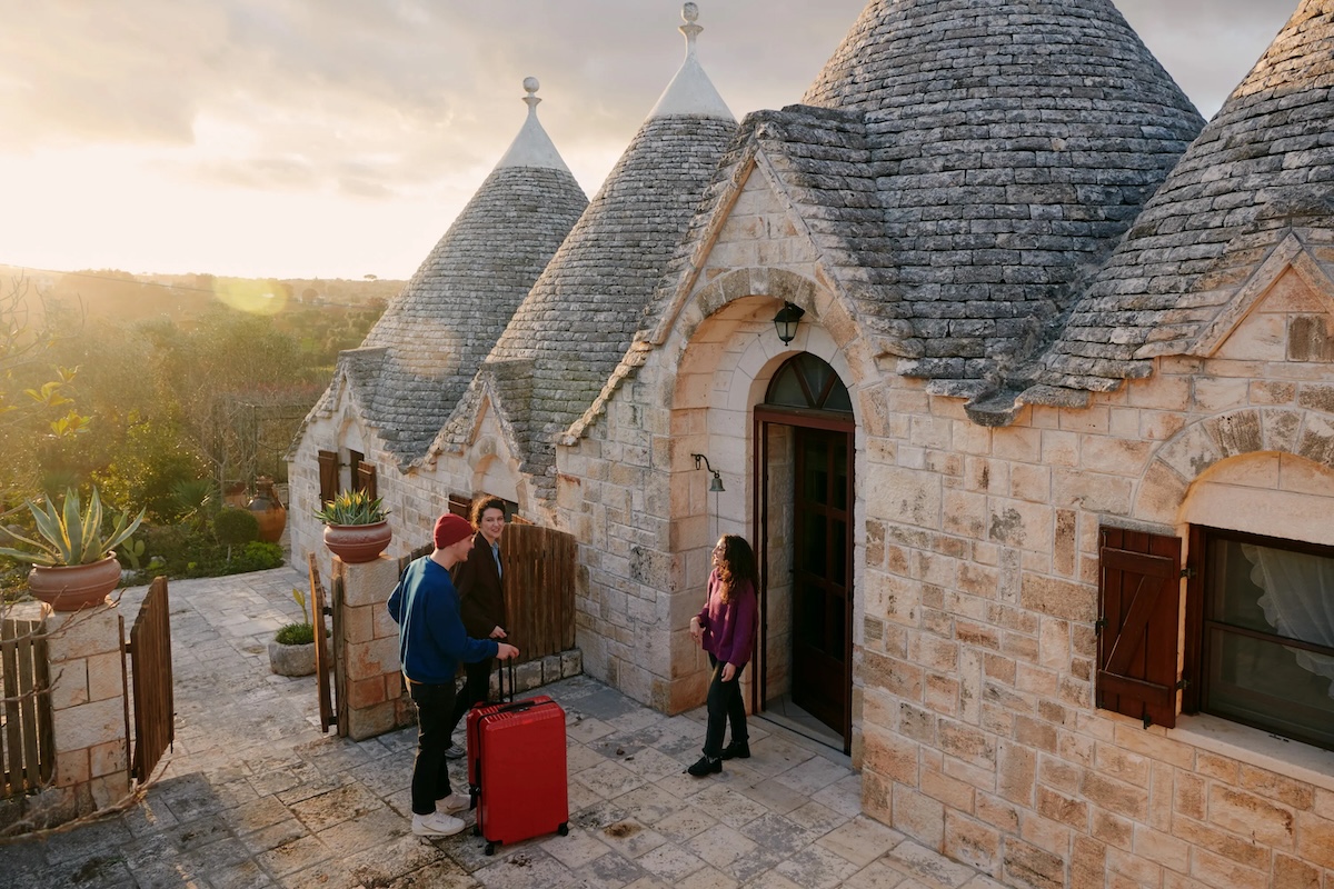 Woman and man with a suitcase greet a woman outside a picturesque home with conical roofs