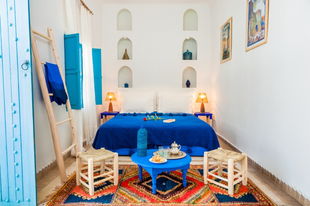 A white bedroom featuring a bed with blue linens and a miniature blue coffee table in front of it, set with tea and cakes for two.