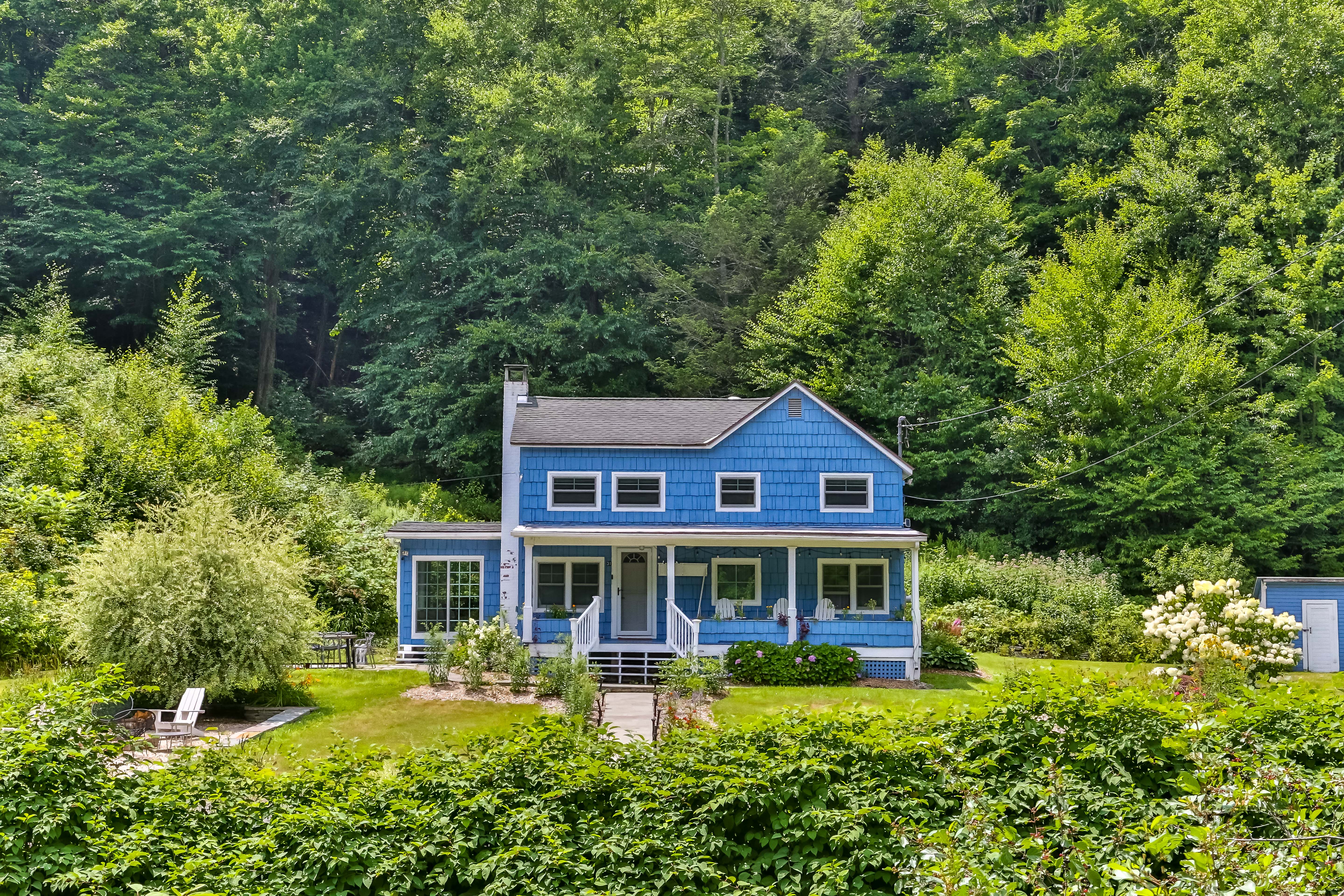 A blue shingled house at the base of a forest with hydrangeas and lush greenery surrounding its large property.