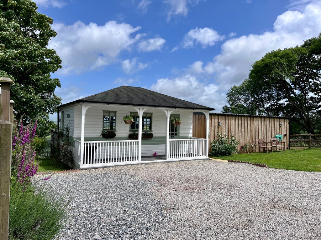 Exterior shot of a timber clad white cabin in Cornwall. Gravel driveway and lawn in the foreground.