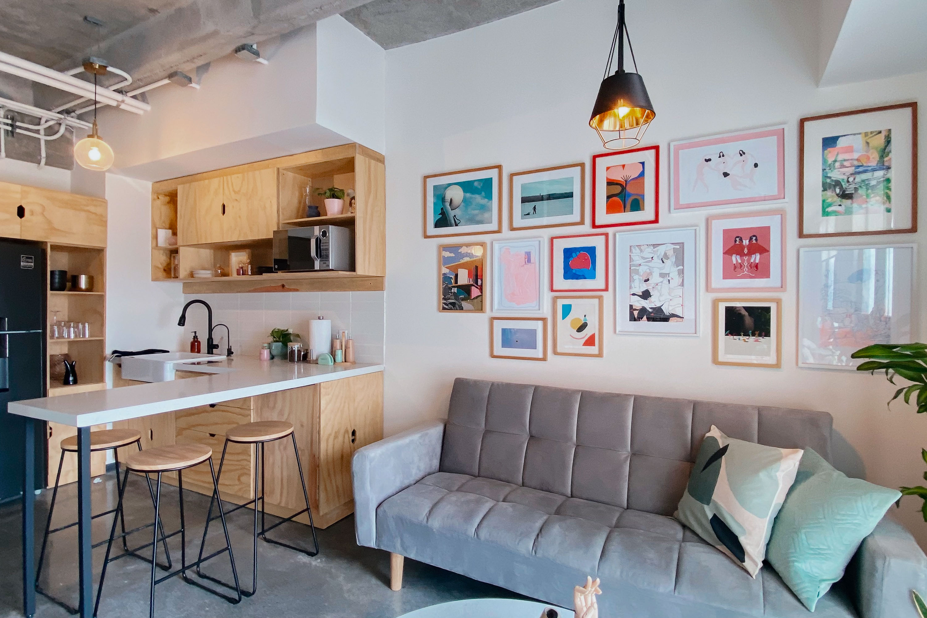 Studio apartment living and kitchen area, with a grey couch and art-filled gallery wall to the right and wooden cabinets and bar stools on the left. 