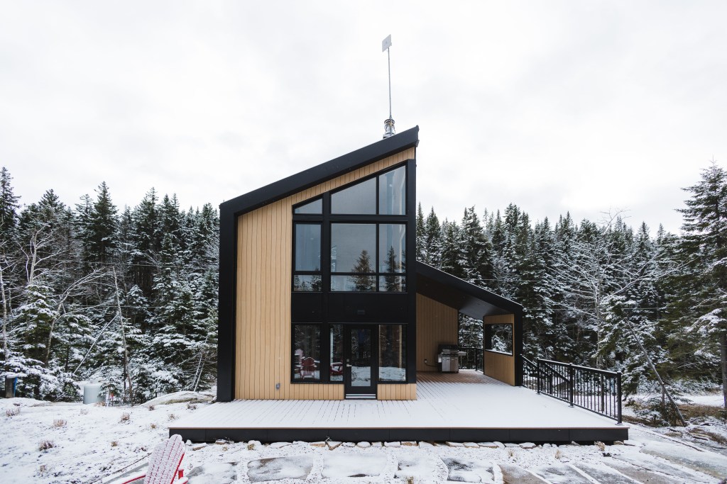 A tiny home with a deck surrounded by forestry in snow. 