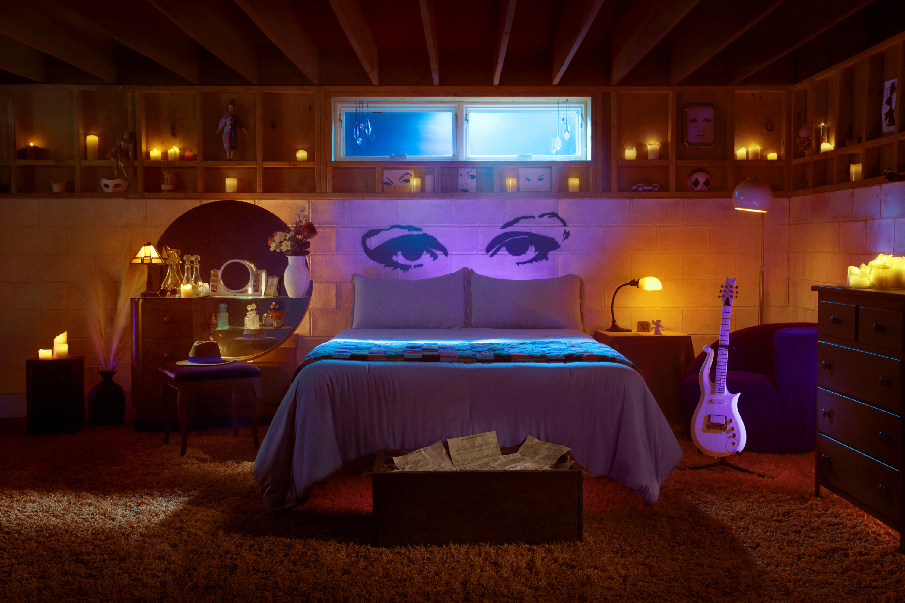 Basement bedroom with a white guitar nearby, backed by spray painted eyes above the pillows. 