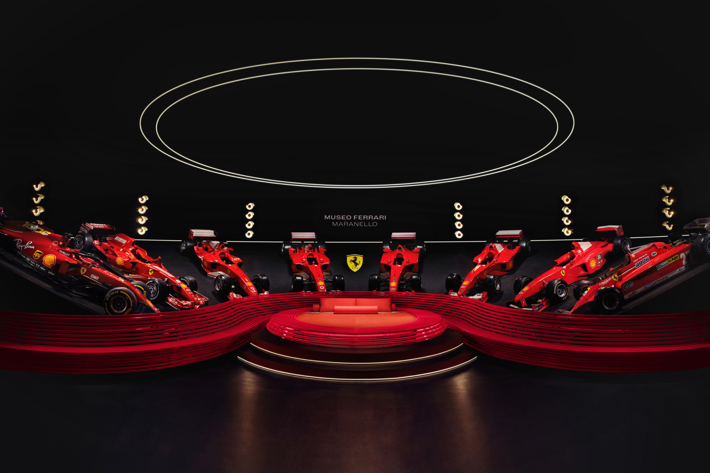 Eight red Ferrari cars on display in a semi circle, framing a circular red bed with a orb light above it.