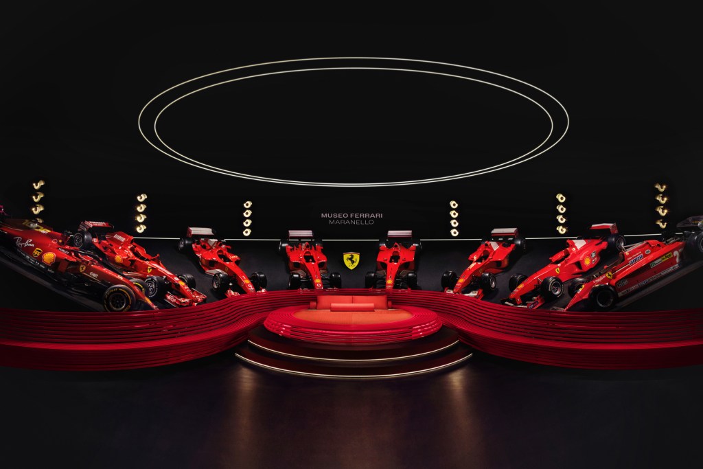 Eight red Ferrari cars on display in a semi circle, framing a circular red bed with a orb light above it. 