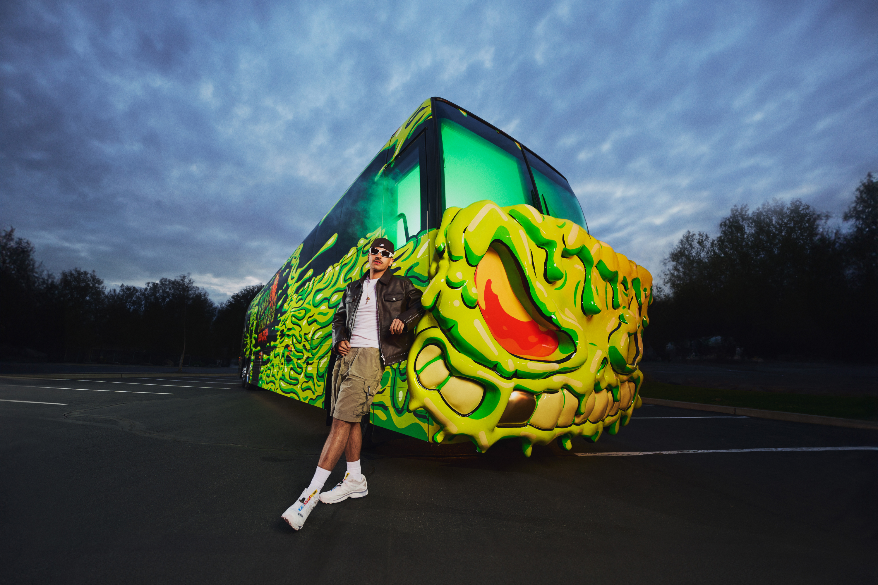 Man leaning up against a loud, green and yellow neon tour bus in an other wise empty parking lot on a cloudy night.