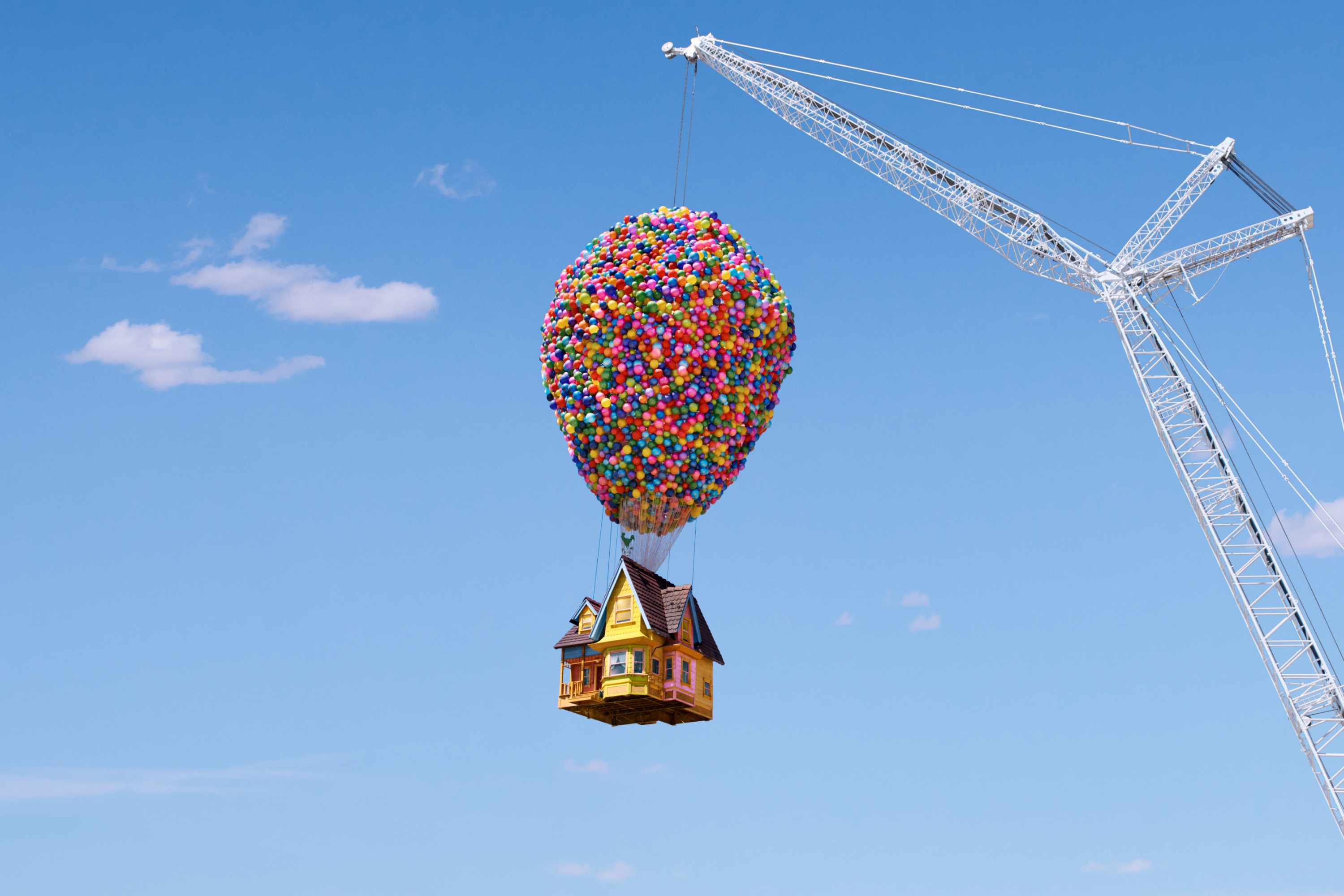 A house suspended by balloons, being lifted by a metal crane, high in a bright blue sky.