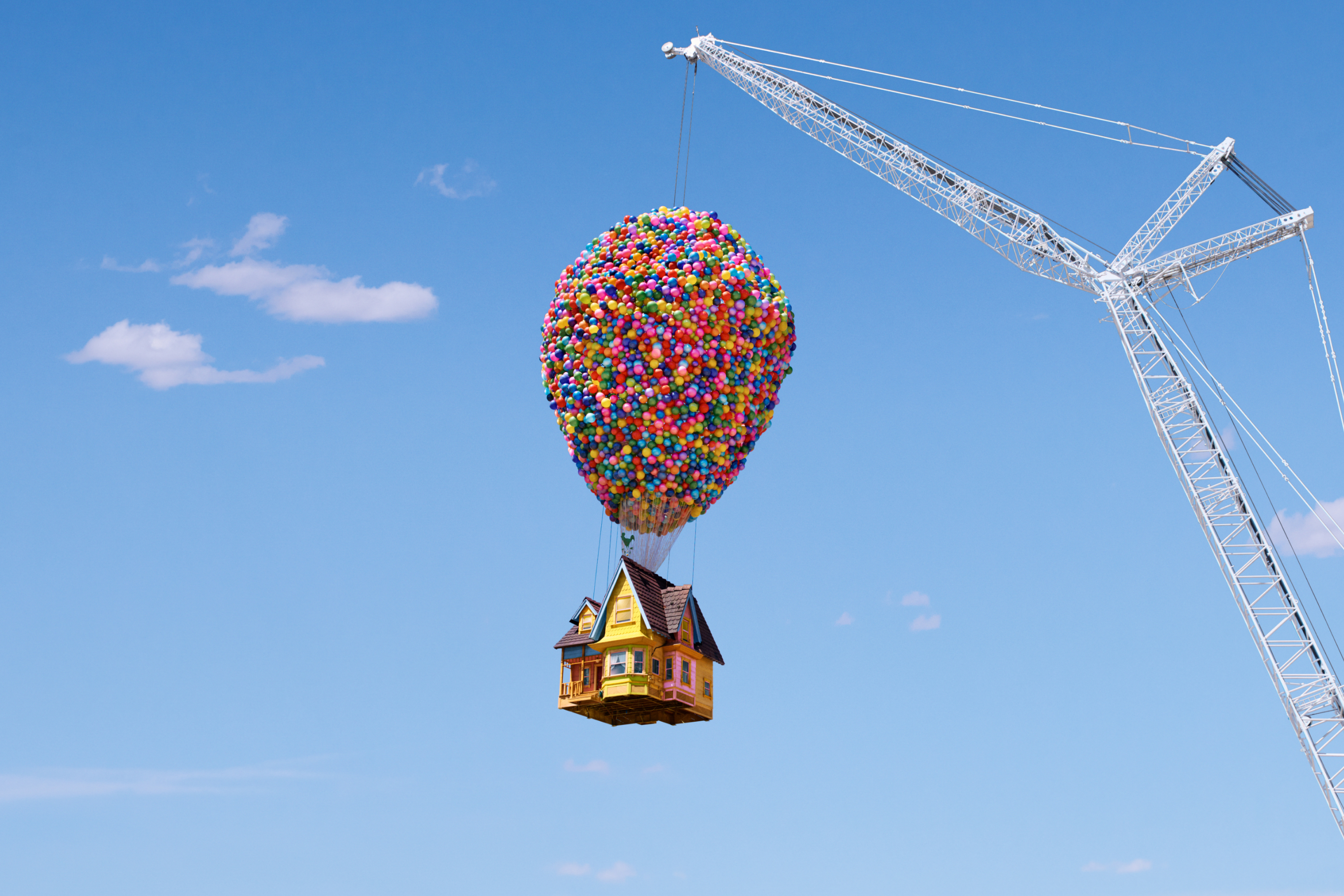A house suspended by balloons, being lifted by a metal crane, high in a bright blue sky. 