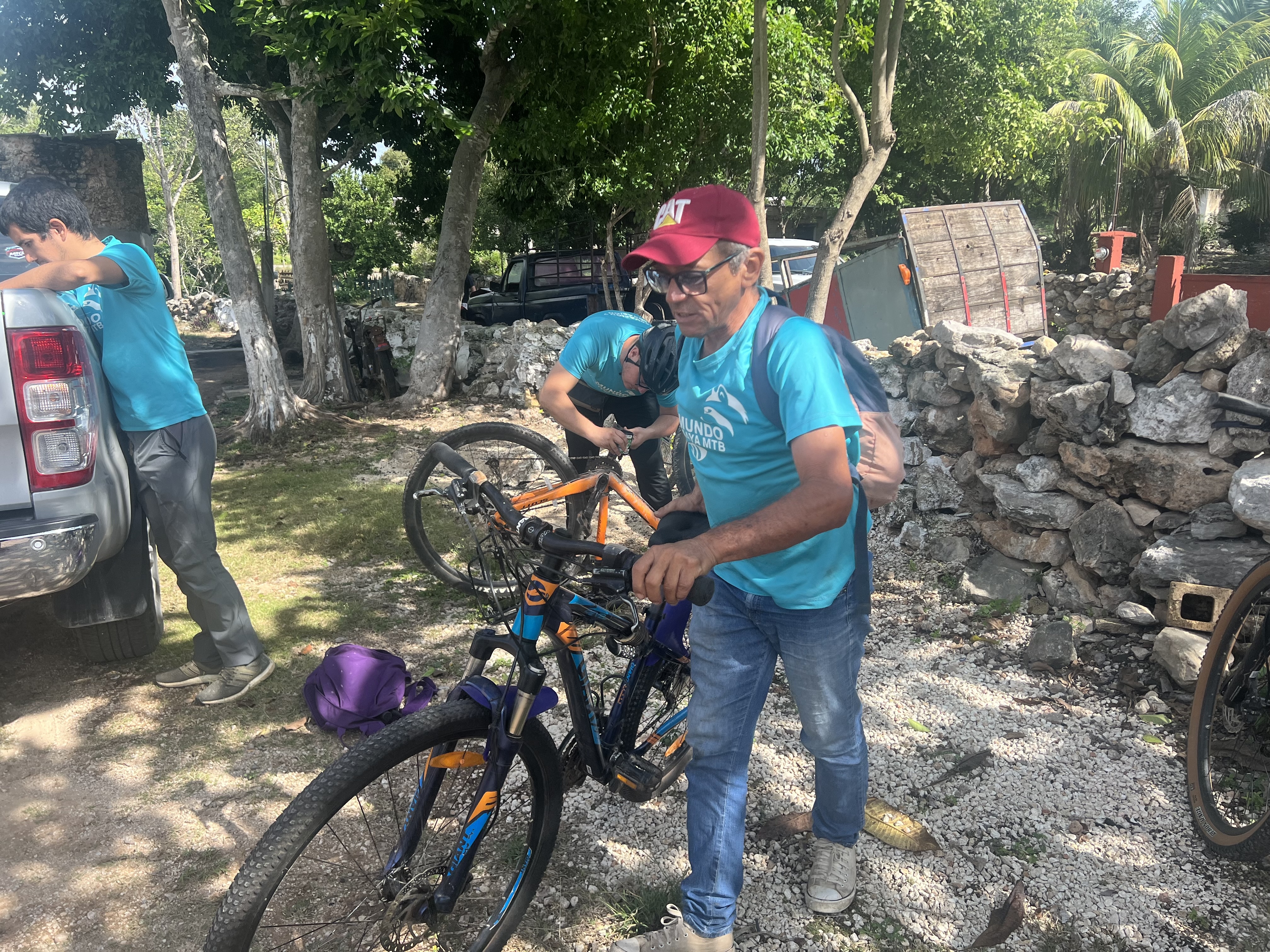 Man in a blue t-shirt and red baseball cap holds a bike while two other men prepare bikes in the background