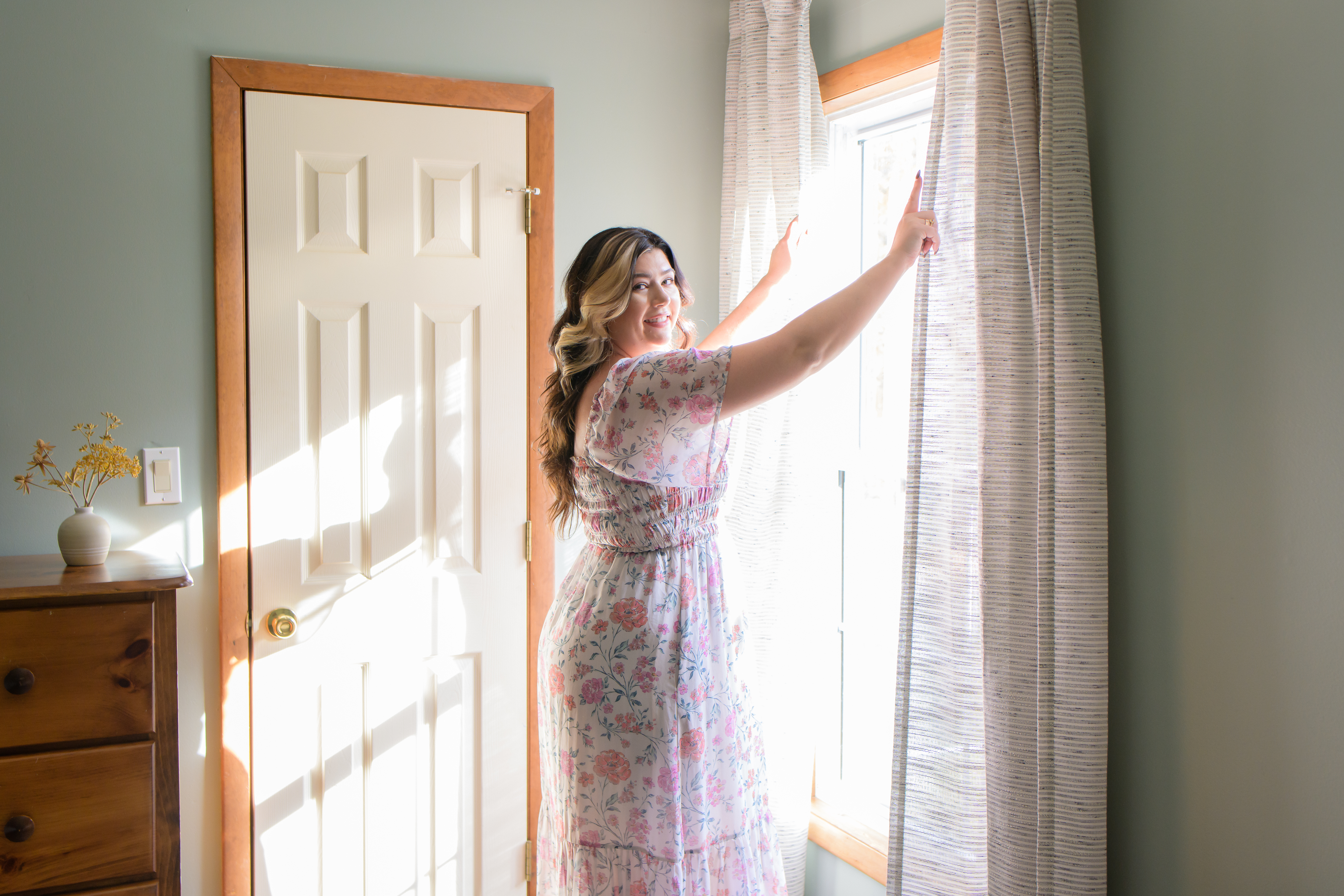 Woman in floral dress stand in front of a window, holding two curtains aside as she looks at the camera