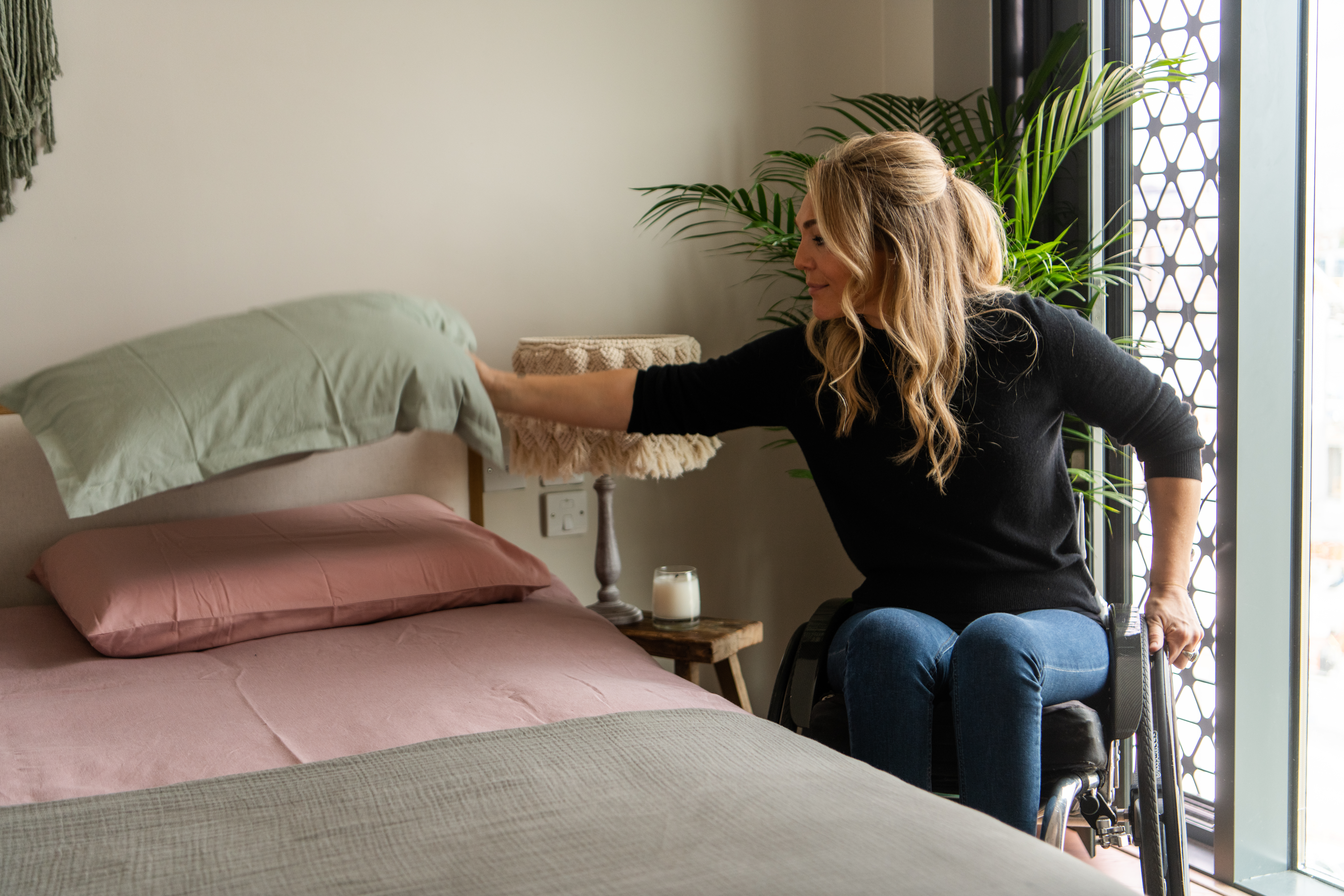 Blonde woman in wheelchair sets a green pillow on a bed as she finishes making the bed.