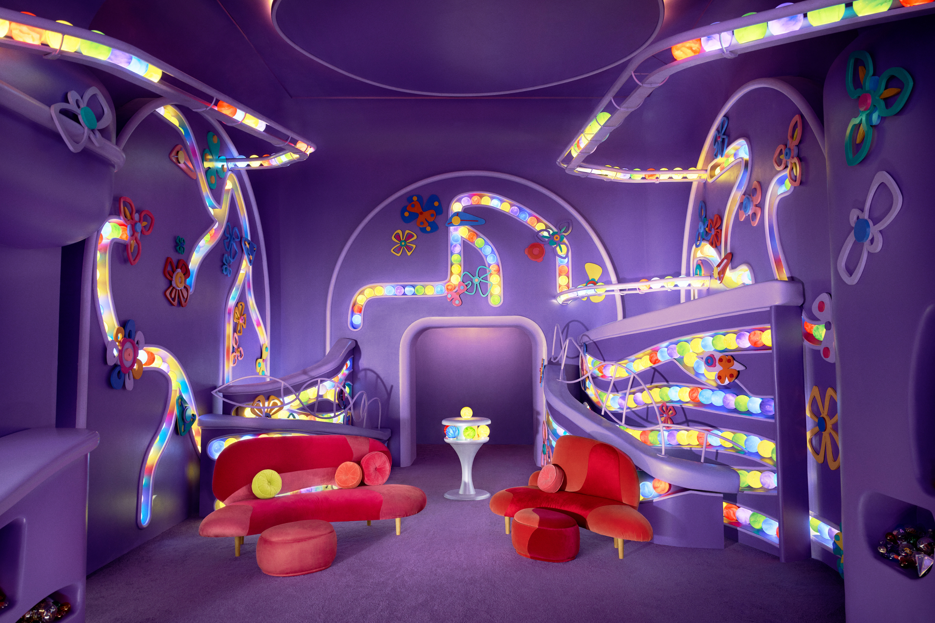 Purple room that has tubes running along the walls filled with green, blue, yellow, purple, and red balls. In the center of the room stand two modern red couches and a pedestal containing the bright colored balls in a ring near the top with a single yellow ball on top.