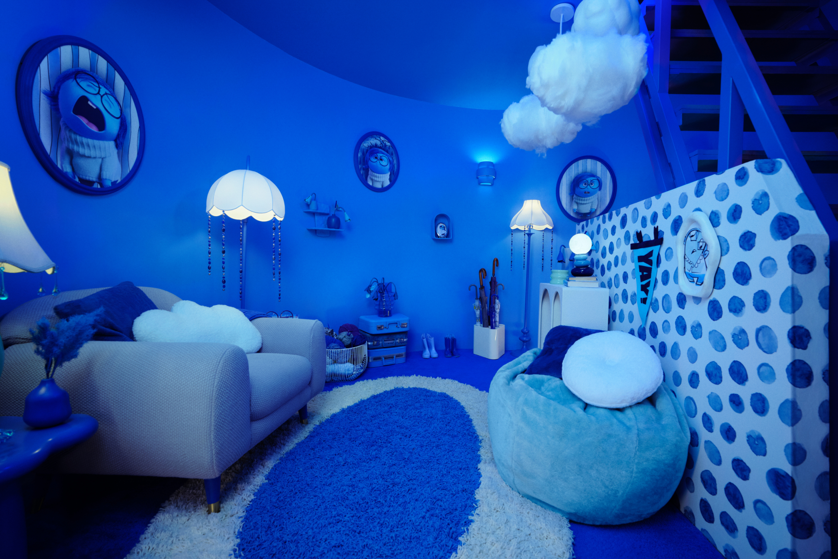 A blue staircase leads down into a windowless dimly lit blue room with a big armchair, blue pouf chairs, an umbrella stand, and some blue cases and boots. There are several portraits of a blue cartoon female looking sad or upset with big glasses, a white sweater, and a short blue bob.