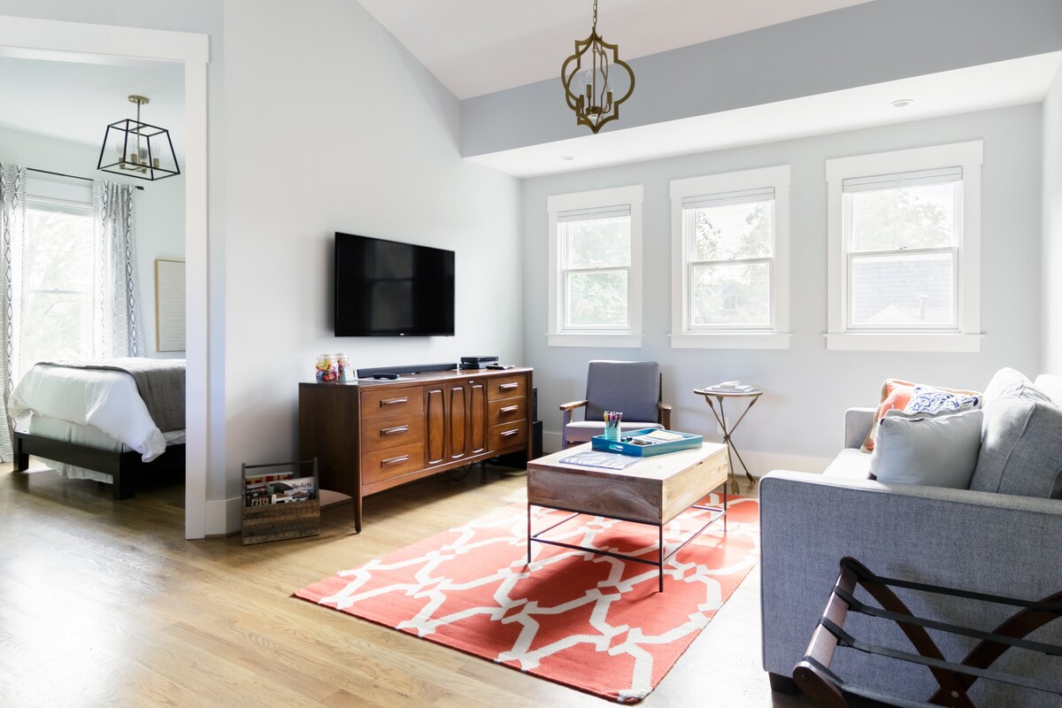 A modern living room with a light gray couch, wooden coffee table on a red and white rug, and a wall-mounted TV above a wooden credenza. An adjacent bedroom is visible through an open doorway.