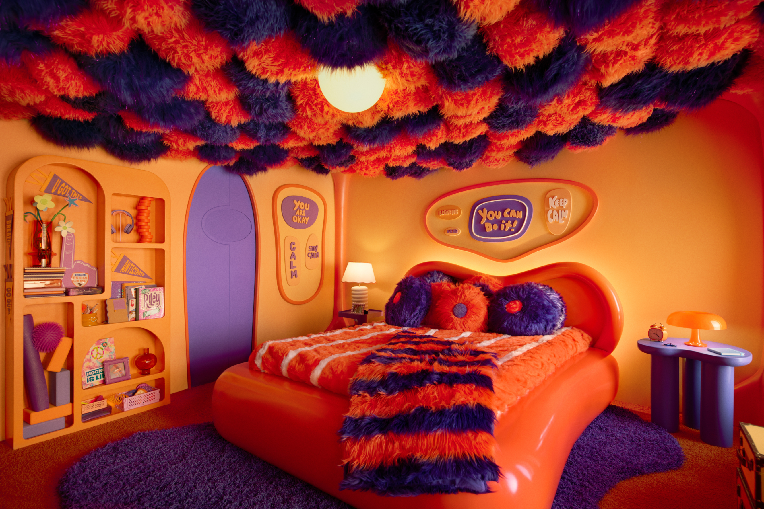 A vivid orange bedroom with a purple rug and door, with a ceiling made of fluffy orange and purple balls. An orange bed is in the center with various knickknacks along the shelves such as headphones, flowers, a yoga mat, a foam finger, books (one of which is titled Riley), and various signs that read "calm," "keep calm," "you are okay," and "you can do it!"