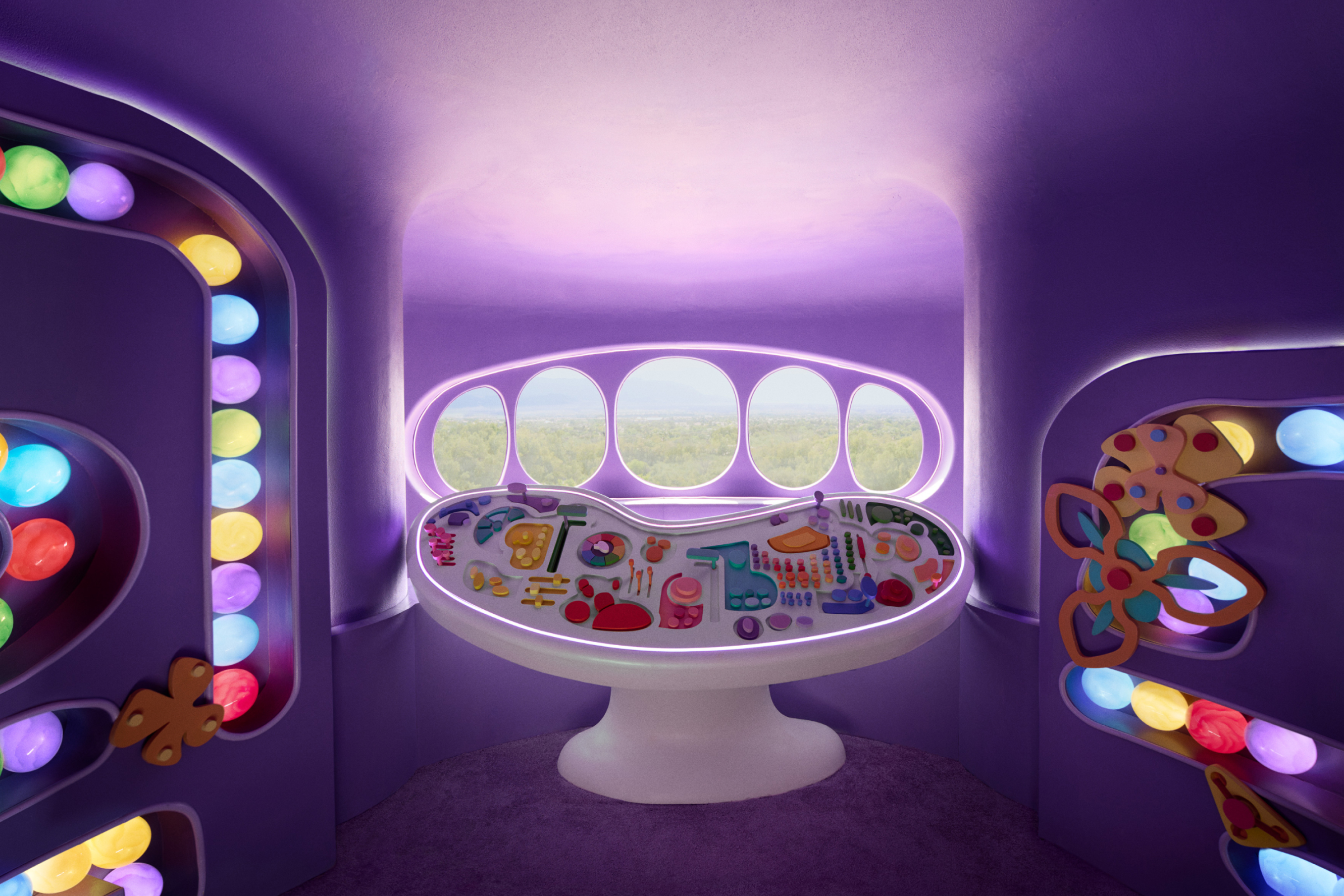 A purple room with bright blue, yellow, red, purple, and green colored balls in tubes along the walls, with a white control panel with various colored knobs and buttons in the center overlooking a forest landscape through five circular windows.