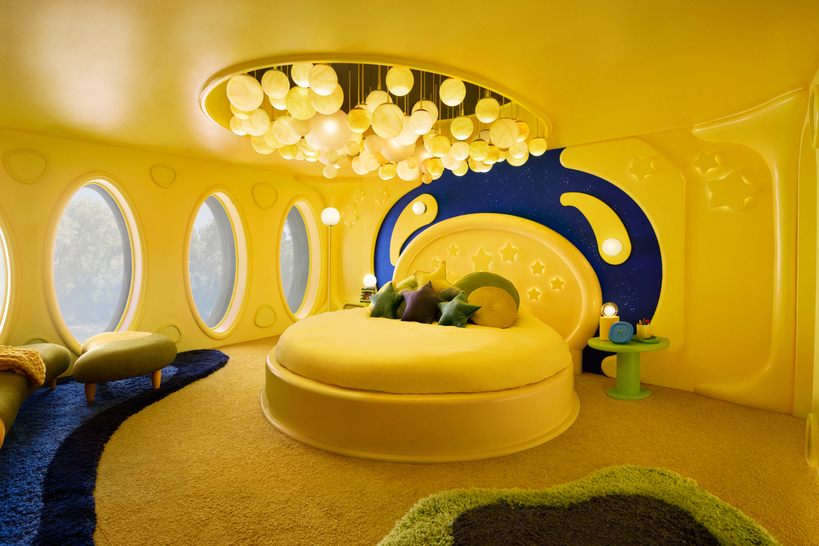 A bright yellow bedroom with a dark blue rug and area on the wall behind the bed that looks like the night sky with abstract yellow shooting stars. A few yellow stars adorn the walls and bed, as well as green, purple, and yellow star-shaped pillows. Dozens of yellow sphere lights hang from a recessed area in the ceiling. There are several large circular windows.