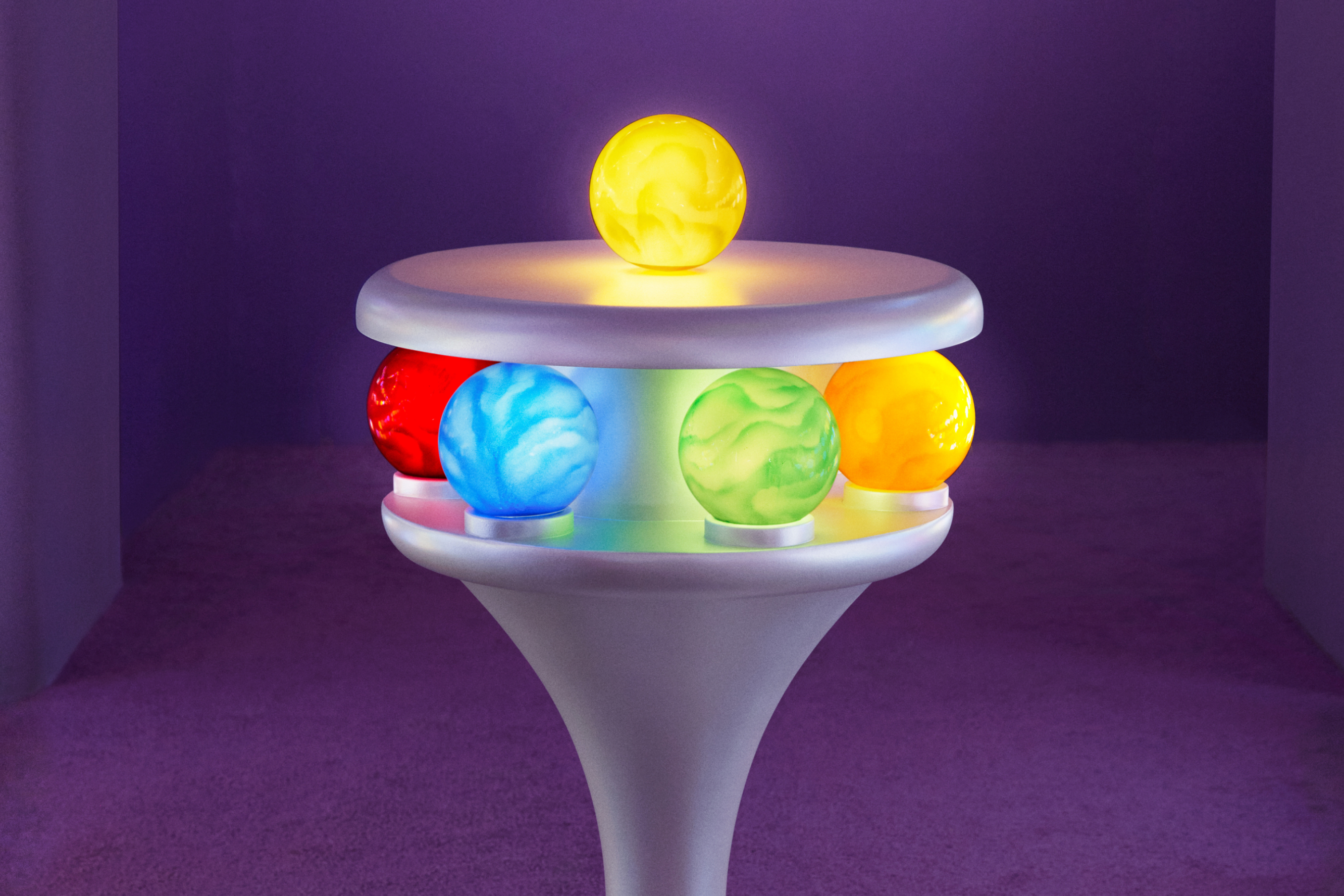 A pedestal in a purple room containing red, blue, green, and yellow balls in a ring near the top with a single yellow ball on top.