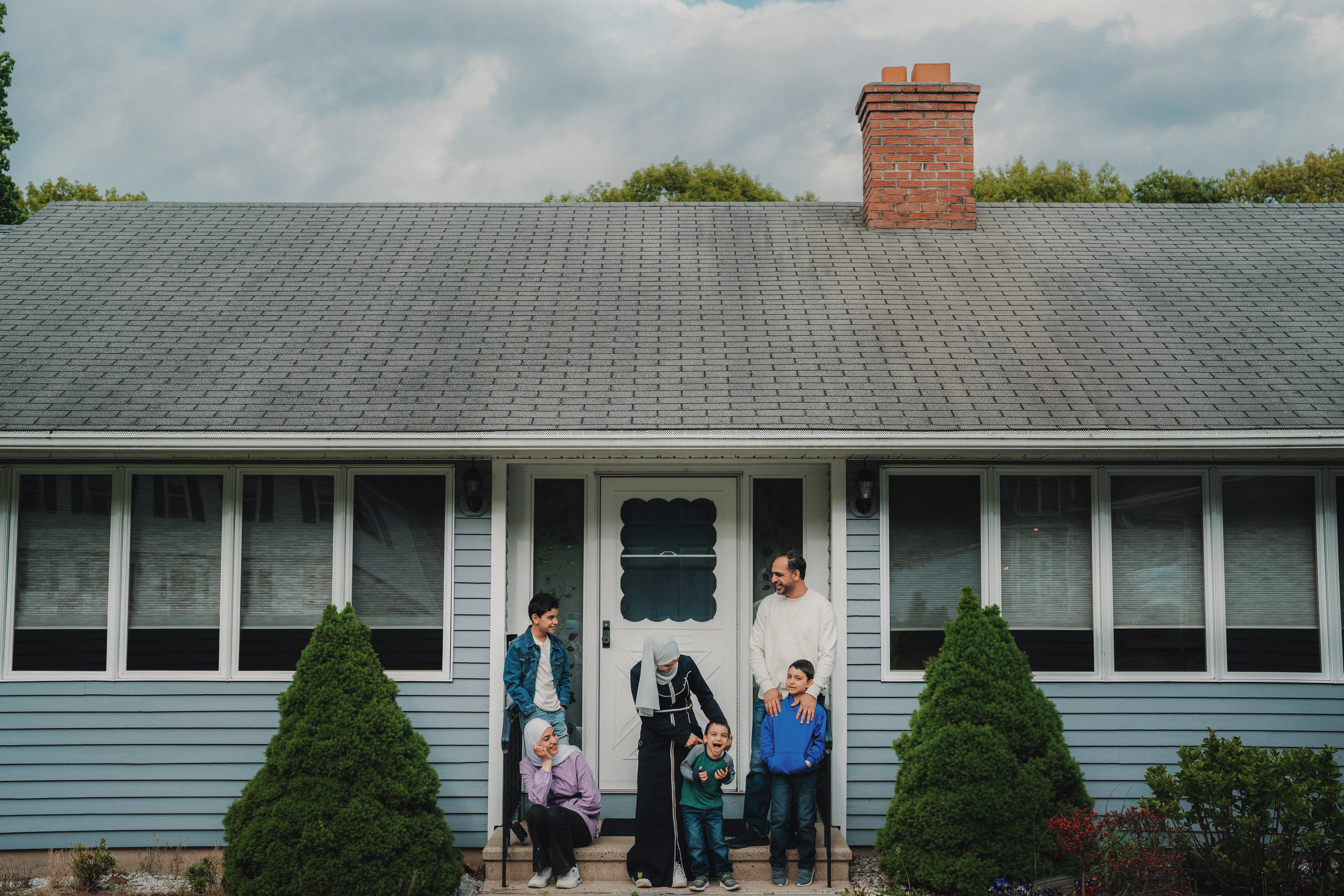 Two parents and their three kids stand on the porch of a house with a grey roof and brick chimney