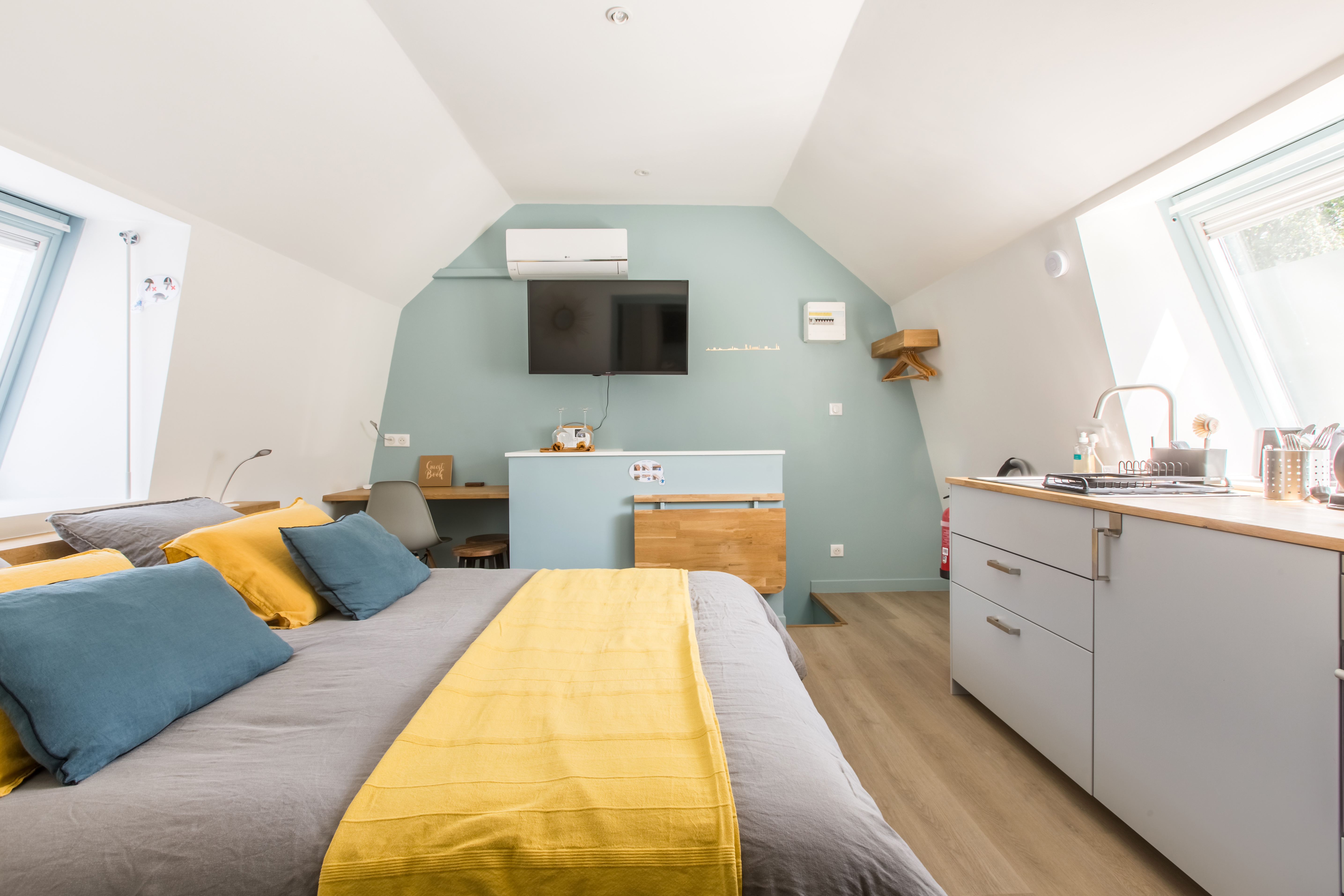 Interior shot of a modern attic studio apartment built under the eaves. Boldly painted aquamarine wall in the background. Bed and kitchenette in the foreground.