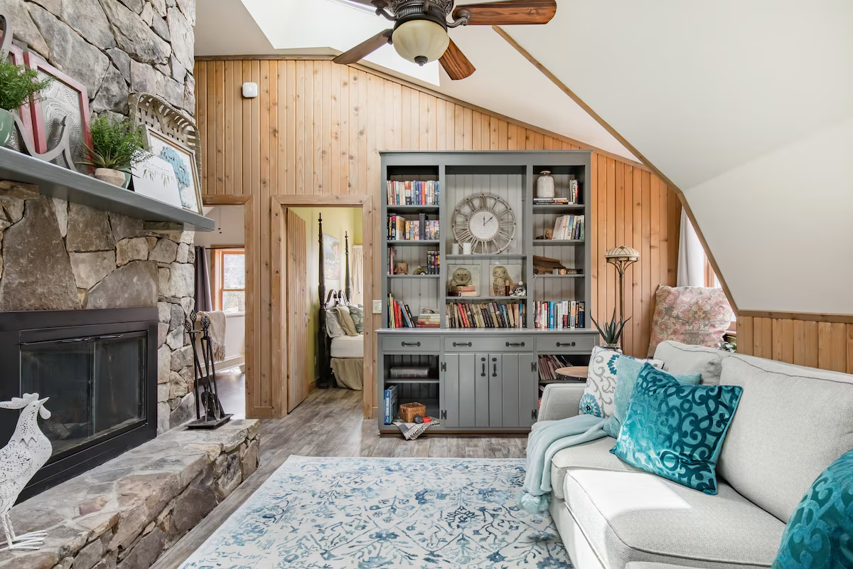 Rustic living room with stone fireplace, wooden wall paneling, built-in bookshelf, white couch with teal pillows, and light blue patterned rug.