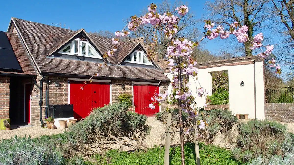Brick building with red doors and white dormer windows, a flowering tree with pink blossoms in the foreground, and a white wall with an open passage next to it.