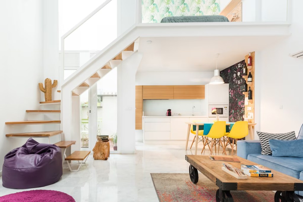 The image portrays a bright, modern living space with an open layout incorporating a kitchen, dining, and living area. The scene includes a staircase on the left leading up to a mezzanine with a patterned leafy wallpaper and a bed.