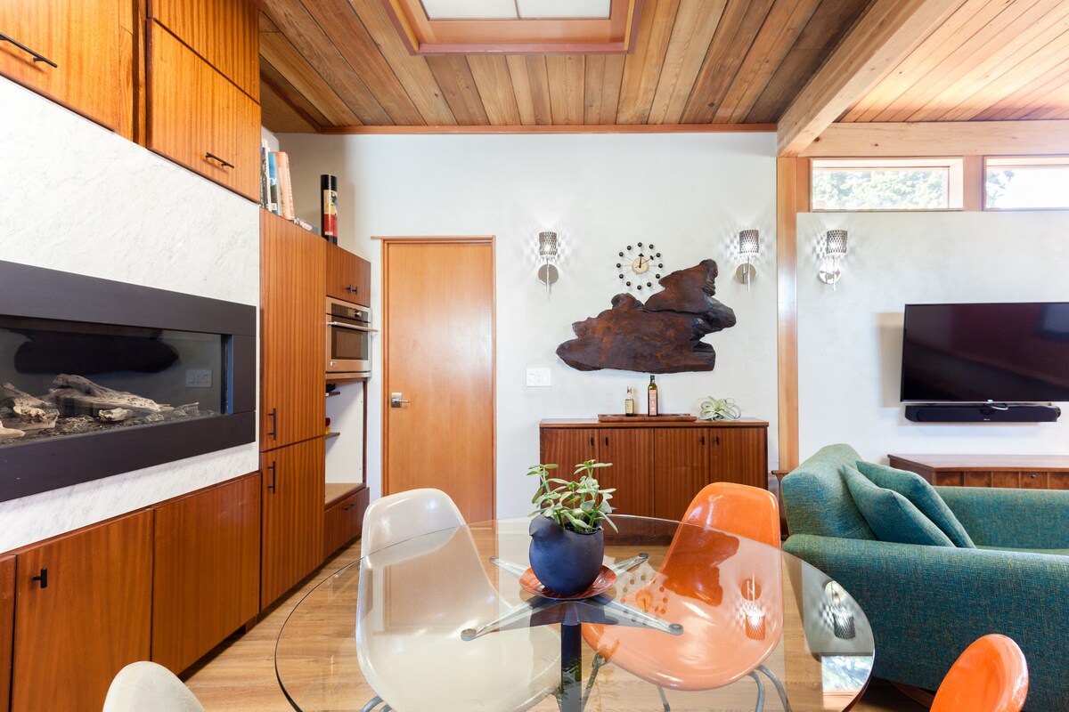 Contemporary living room with wooden ceiling, skylight, black fireplace in a marble wall, wooden cabinetry, glass dining table with white and orange chairs, teal armchair, wall-mounted television, and a wooden sculpture above a sideboard.