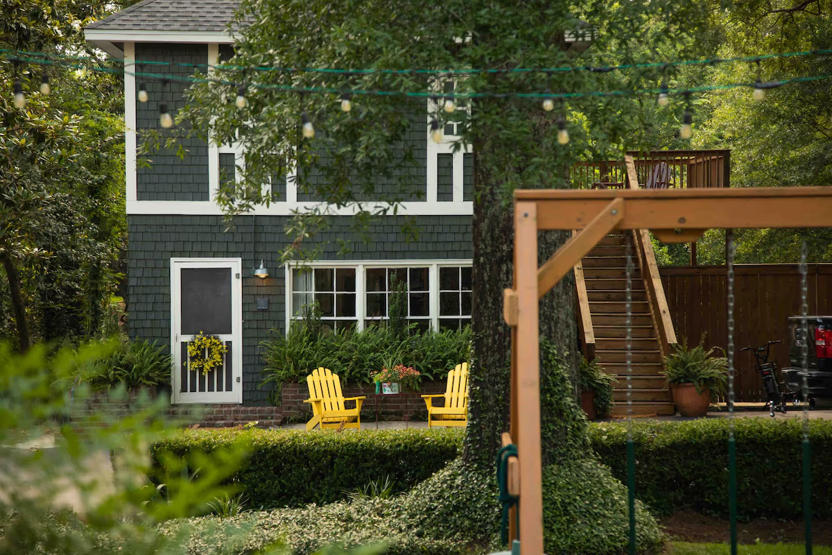 Two-story house with dark green shingles, yellow Adirondack chairs on a patio, and a wooden playset in the foreground.