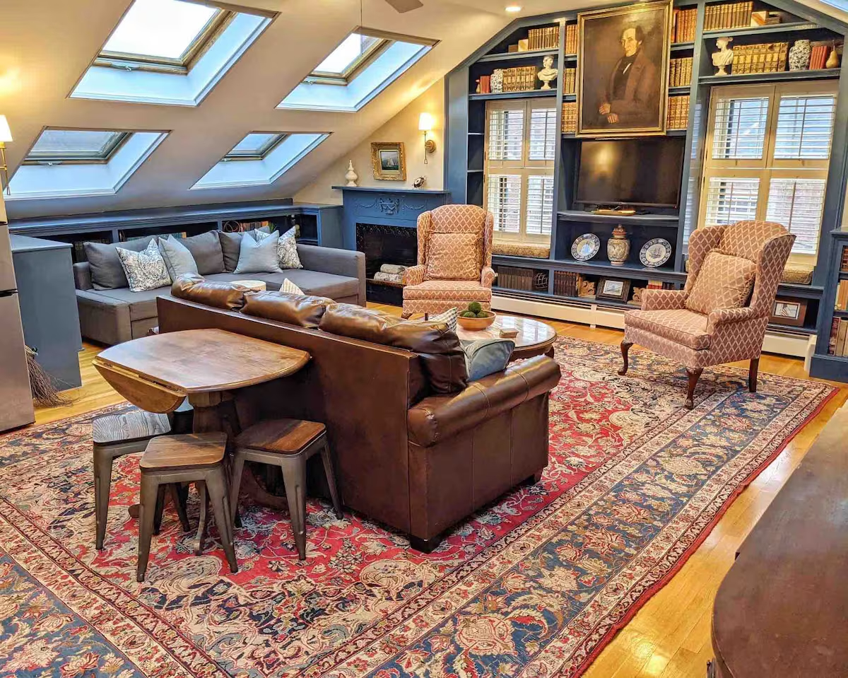 cozy, well-lit living room with a mix of traditional and contemporary decor. The space features four skylights on the slanted ceiling, filling the room with natural light. Below the skylights is a large sectional gray couch adorned with decorative pillows. Directly in front of the couch is a polished wooden coffee table and two patterned armchairs. The seating area is positioned on a large, ornate oriental rug with red, blue, and beige floral designs.