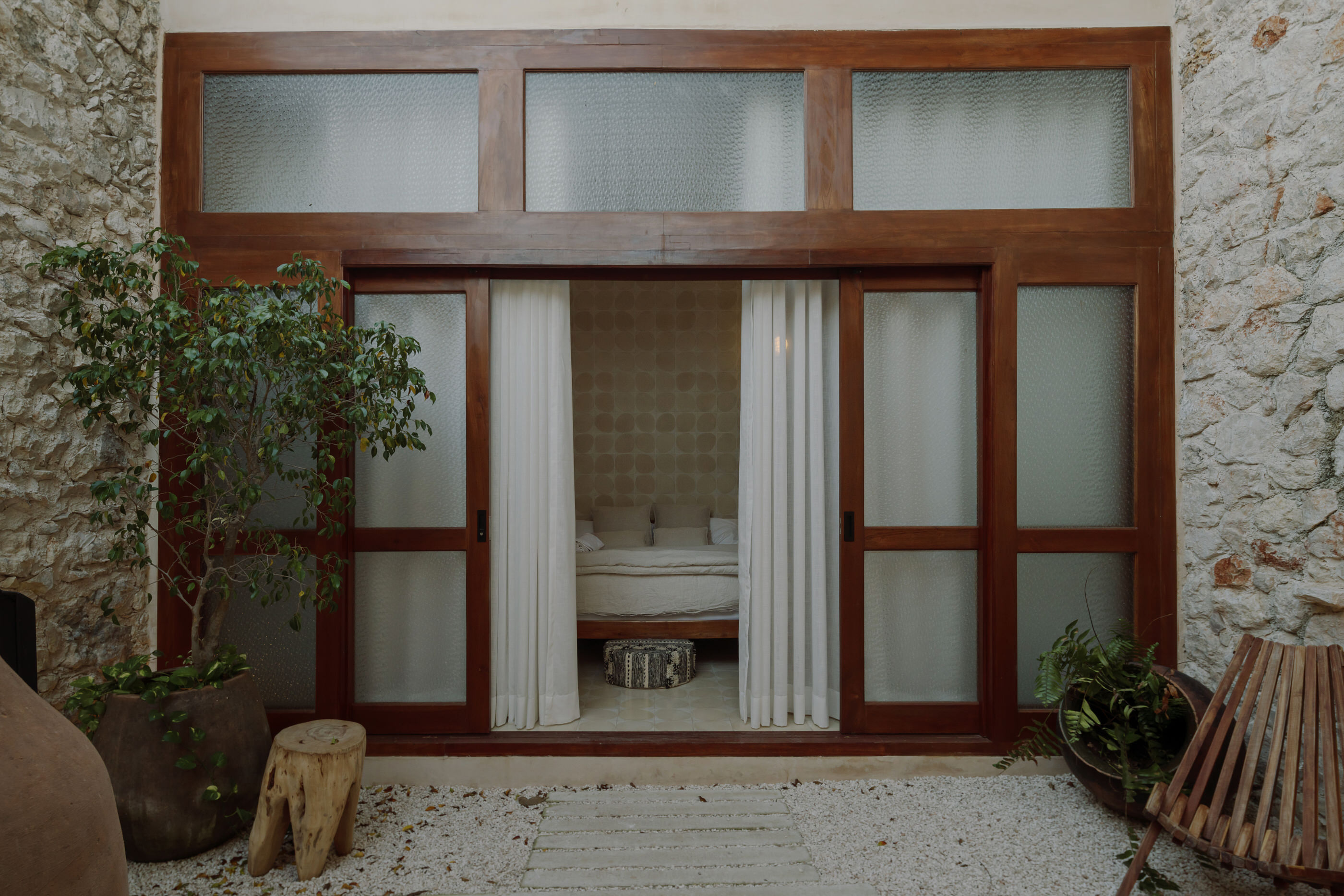 Front view of a glass door with view to the interior of a bedroom with a tree and a chair to the left