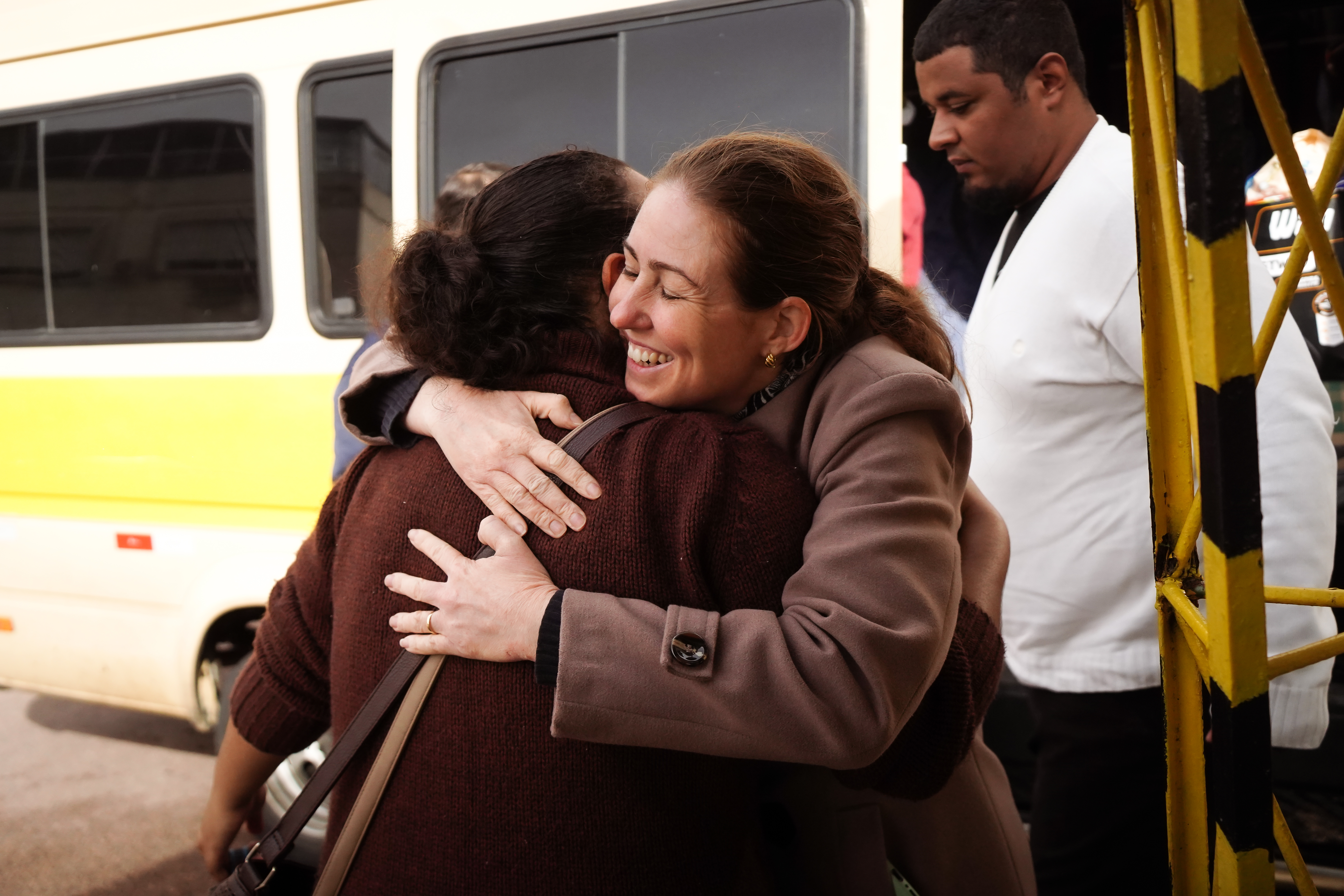 A brown-haired woman smiles as she embraces a woman in a brown sweater in front of a bus from which more people emerge
