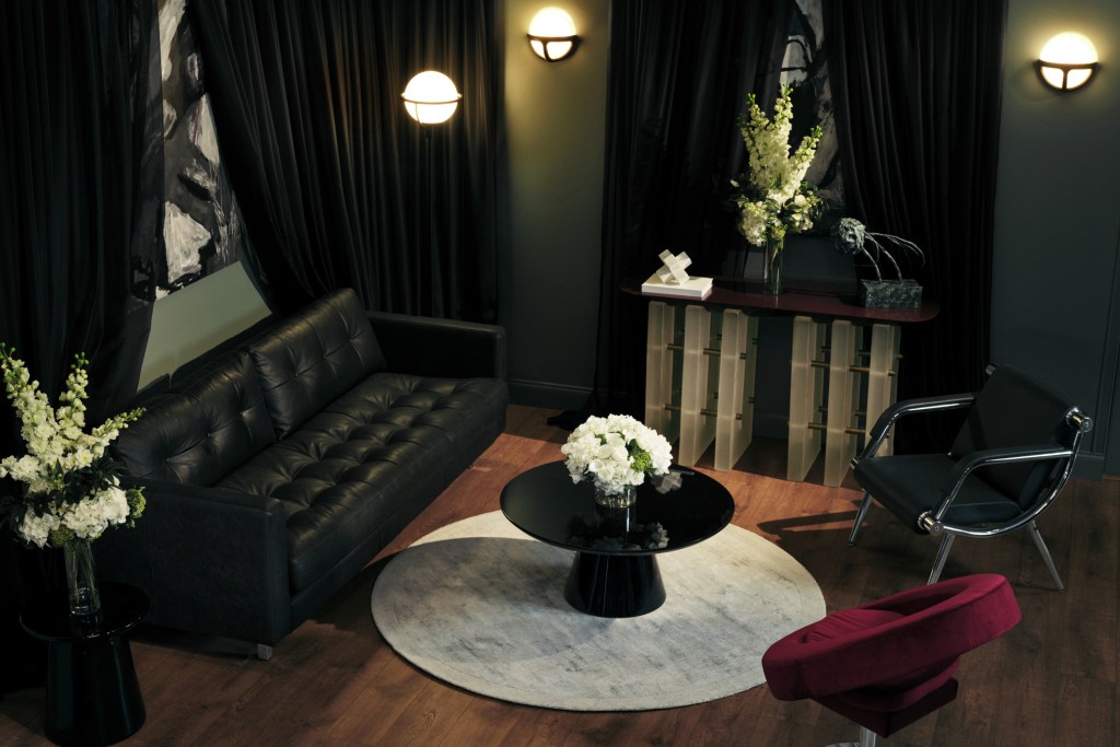 Living room setting with black leather couch, black circular coffee table with a vase of white flowers and two mismatching chairs with green walls and sconce lighting.