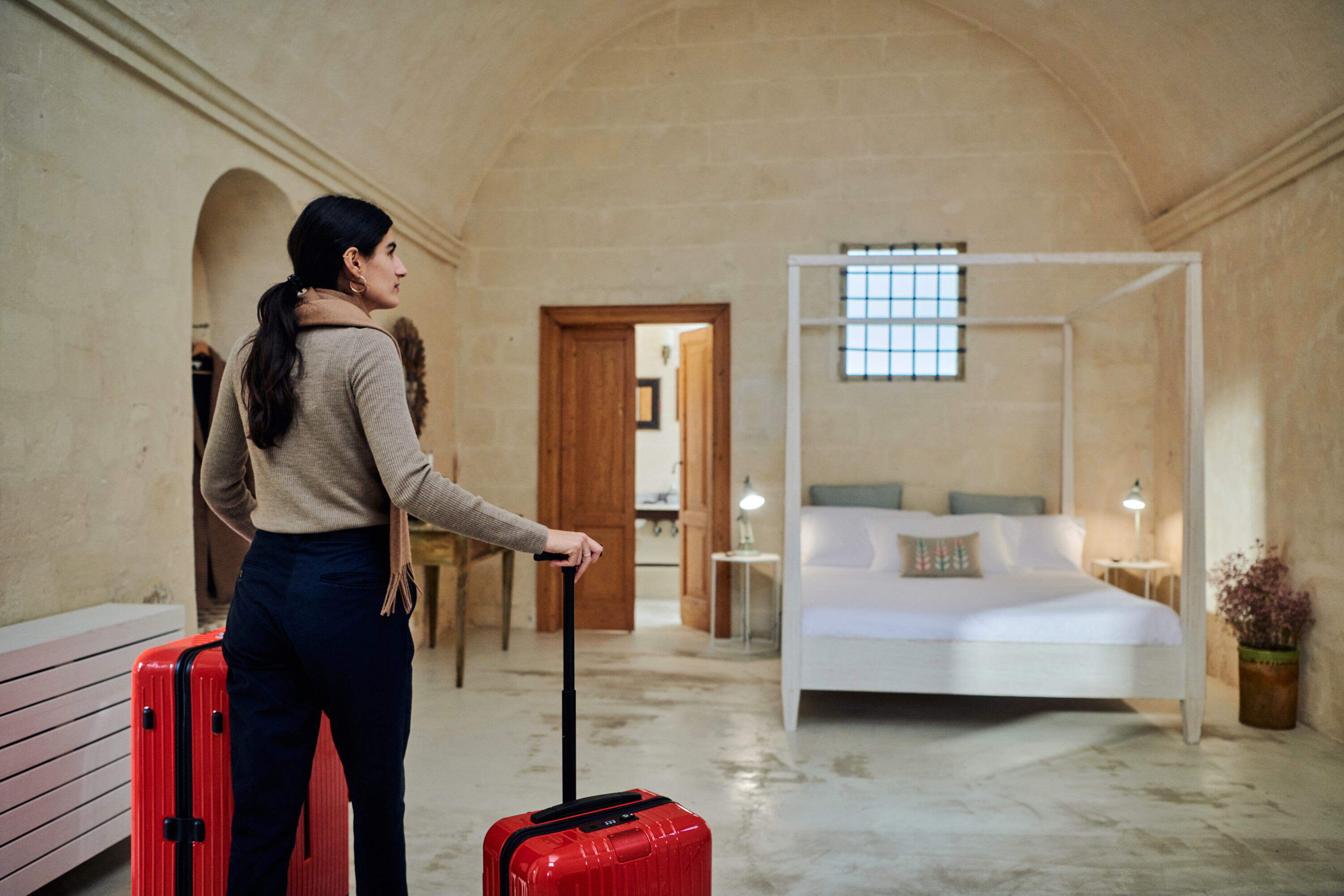 A woman with a red suitcase in a stone-walled bedroom with a canopy bed.