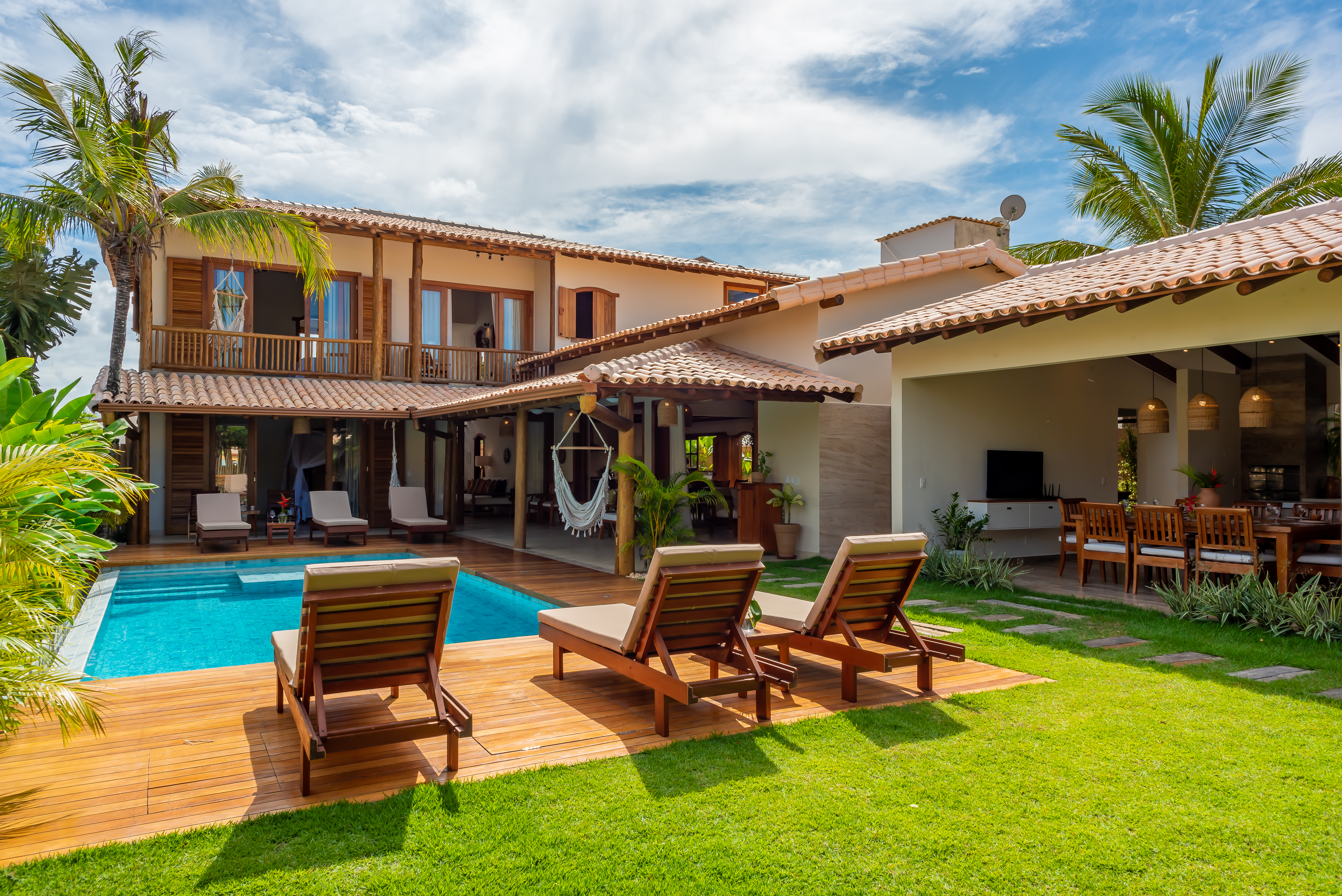 An outer view of a modern villa with a pool surrounded by wooden armchairs, lush green grass and palm trees. The villa has a tile roof, large windows and a spanish courtyard with a hanging bed. The interior is visible through open doors, displaying a cozy living and dining space.