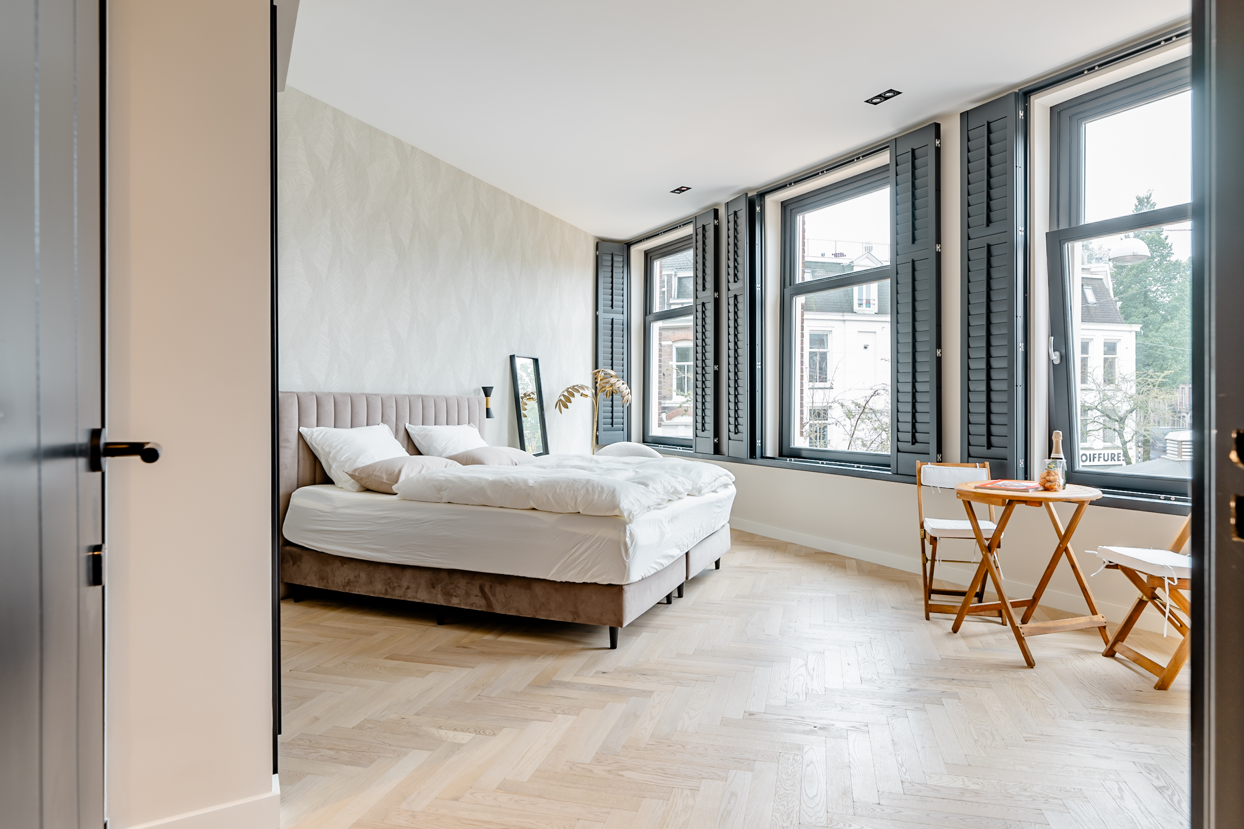 A serene bedroom with herringbone floors, an upholstered beige bed and neutral patterned wallpaper. There are three large windows with turquoise shutters and a table for two in the corner prefect for morning coffee.