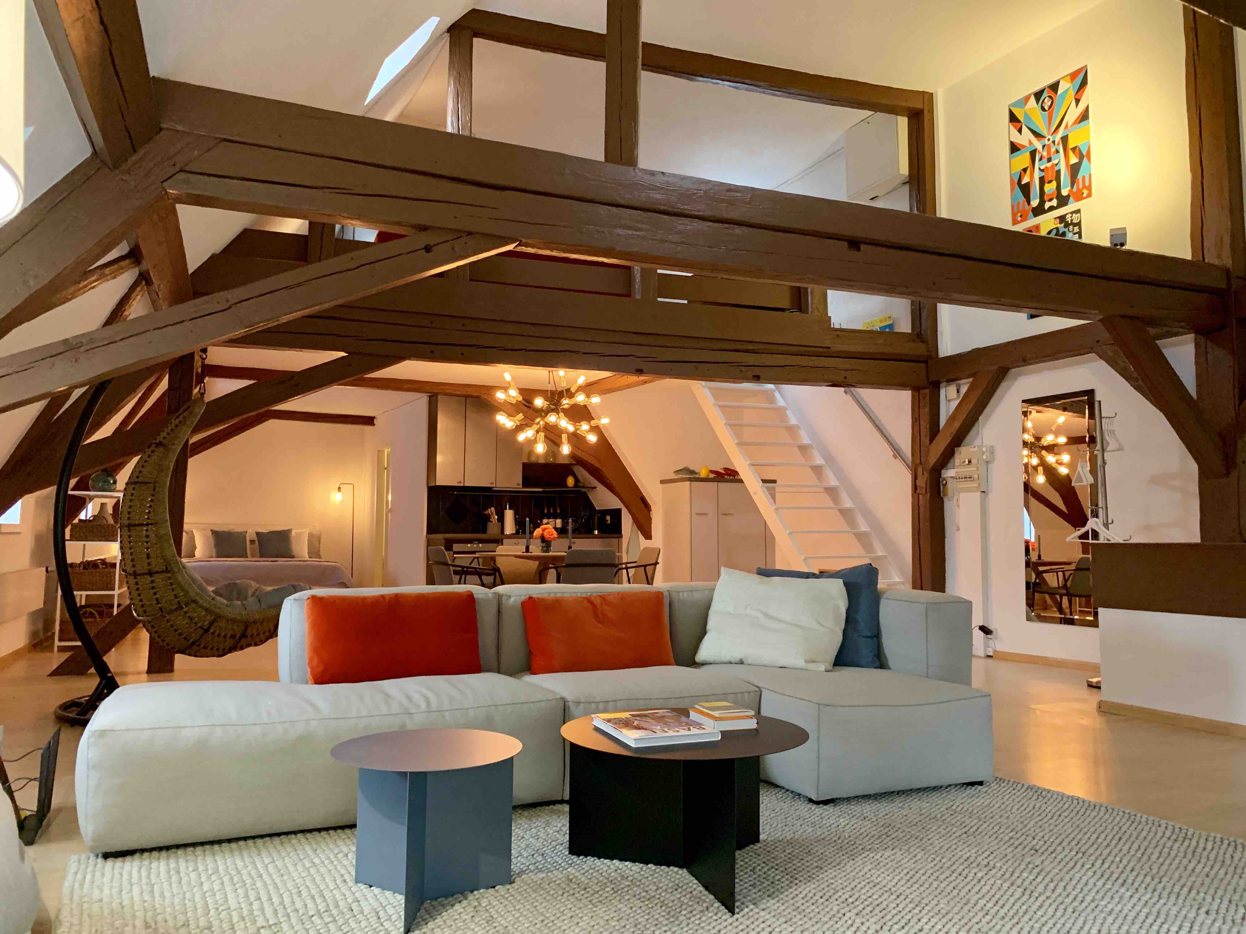 Spacious and modern loft-style living area featuring a cozy sectional sofa with orange and blue accent pillows, a circular black coffee table, and a textured area rug. In the background, a stylish dining area with a contemporary chandelier and a well-equipped kitchen is visible. A hanging wicker chair adds a touch of relaxation, while a staircase leads to a lofted sleeping area, creating an inviting and artistic atmosphere with wooden beams and vibrant wall art.