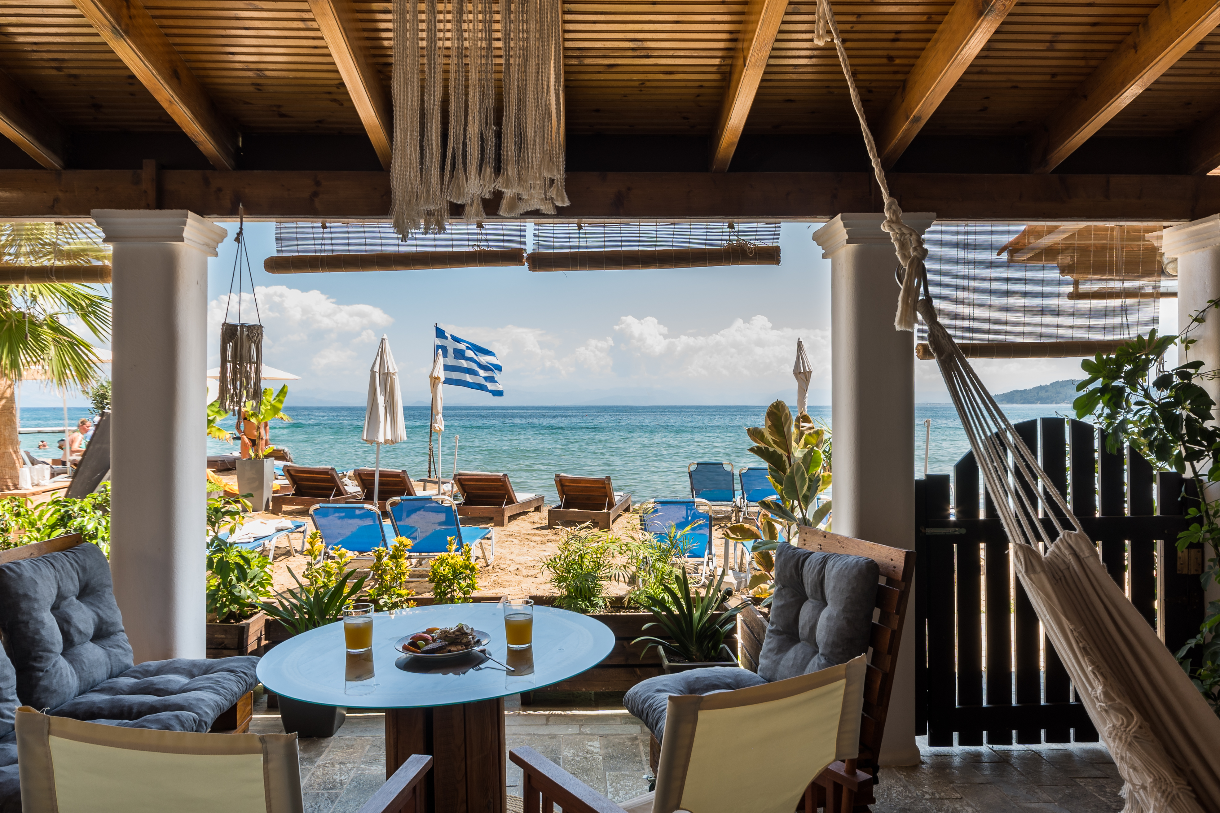 A cozy patio with a view of the beach. The patio is adorned with tropical plants and a hammock, while a Greek flag flutters in the breeze. Beach loungers are set up on the sandy shore, and the deep blue sea is visible in the background under a partly cloudy sky. On the table, there are two glasses of drinks and a plate of food, inviting relaxation and enjoyment in a picturesque setting.