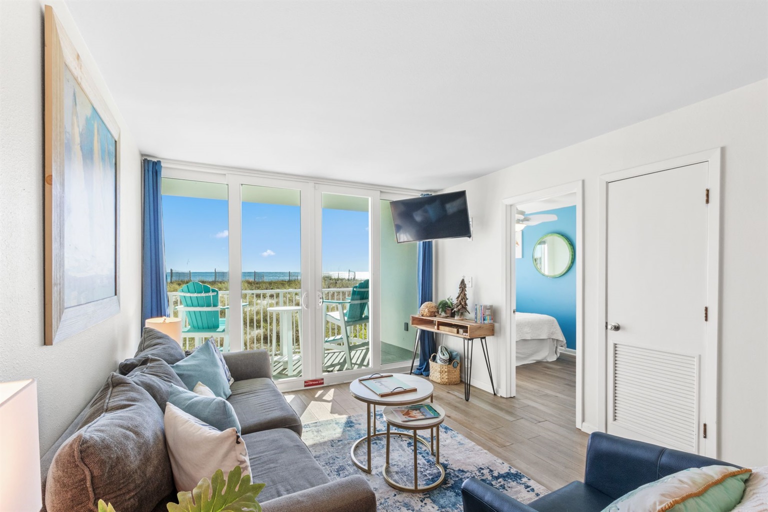 A cozy living room featuring a gray sofa adorned with decorative pillows, a small round coffee table, and a television mounted on the wall. Large glass doors lead to a balcony with a view of the beach and turquoise chairs. The room is bright and airy with light-colored walls and wooden flooring, creating a welcoming seaside ambiance.