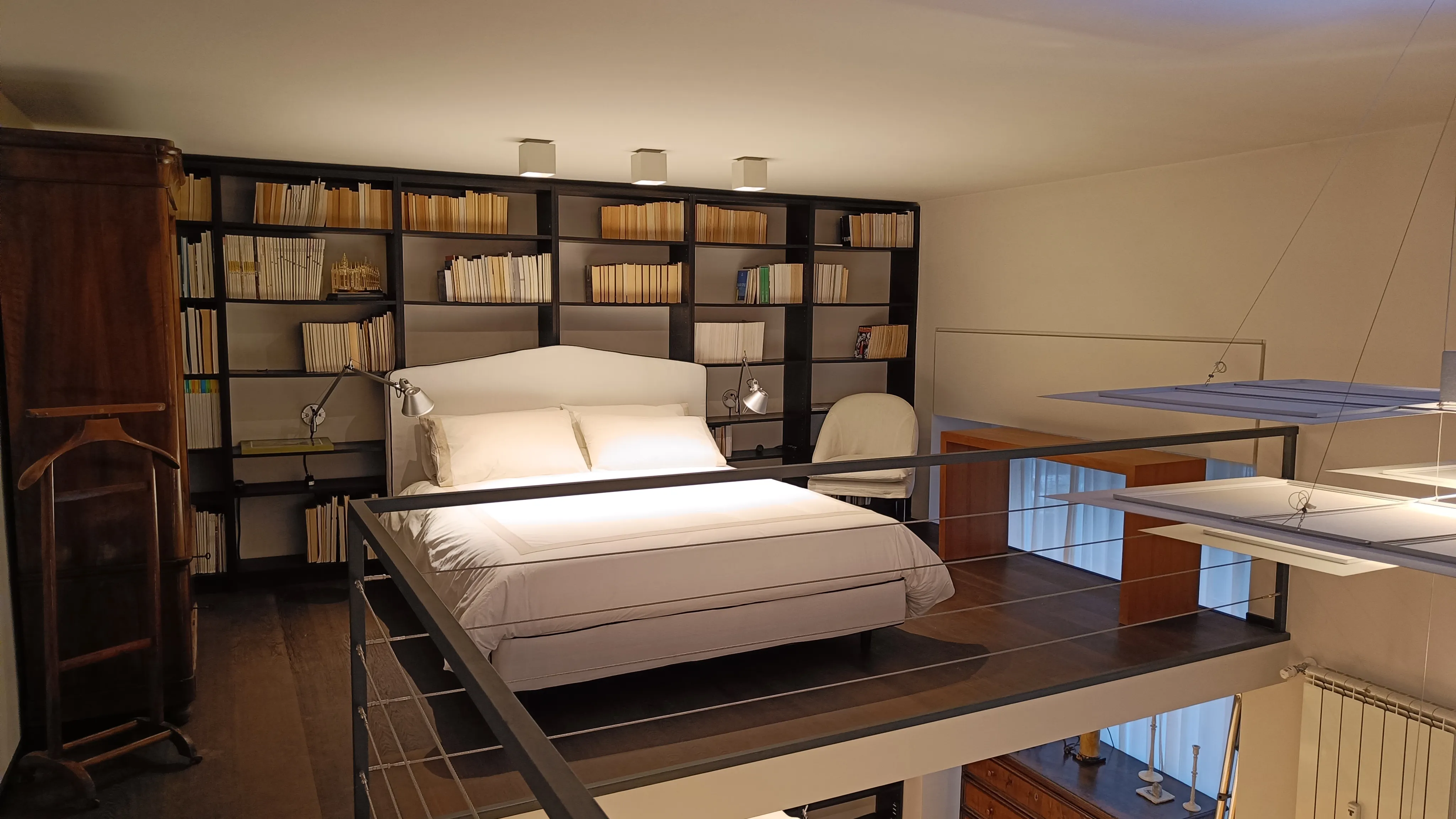 A stylish loft featuring a cozy elevated bed with white linens, surrounded by an extensive bookshelf filled with books. A sleek reading lamp is positioned next to the bed, and a white chair is placed nearby. The wooden wardrobe adds a touch of warmth to the modern decor. The space is well-lit with soft lighting, creating an inviting atmosphere.